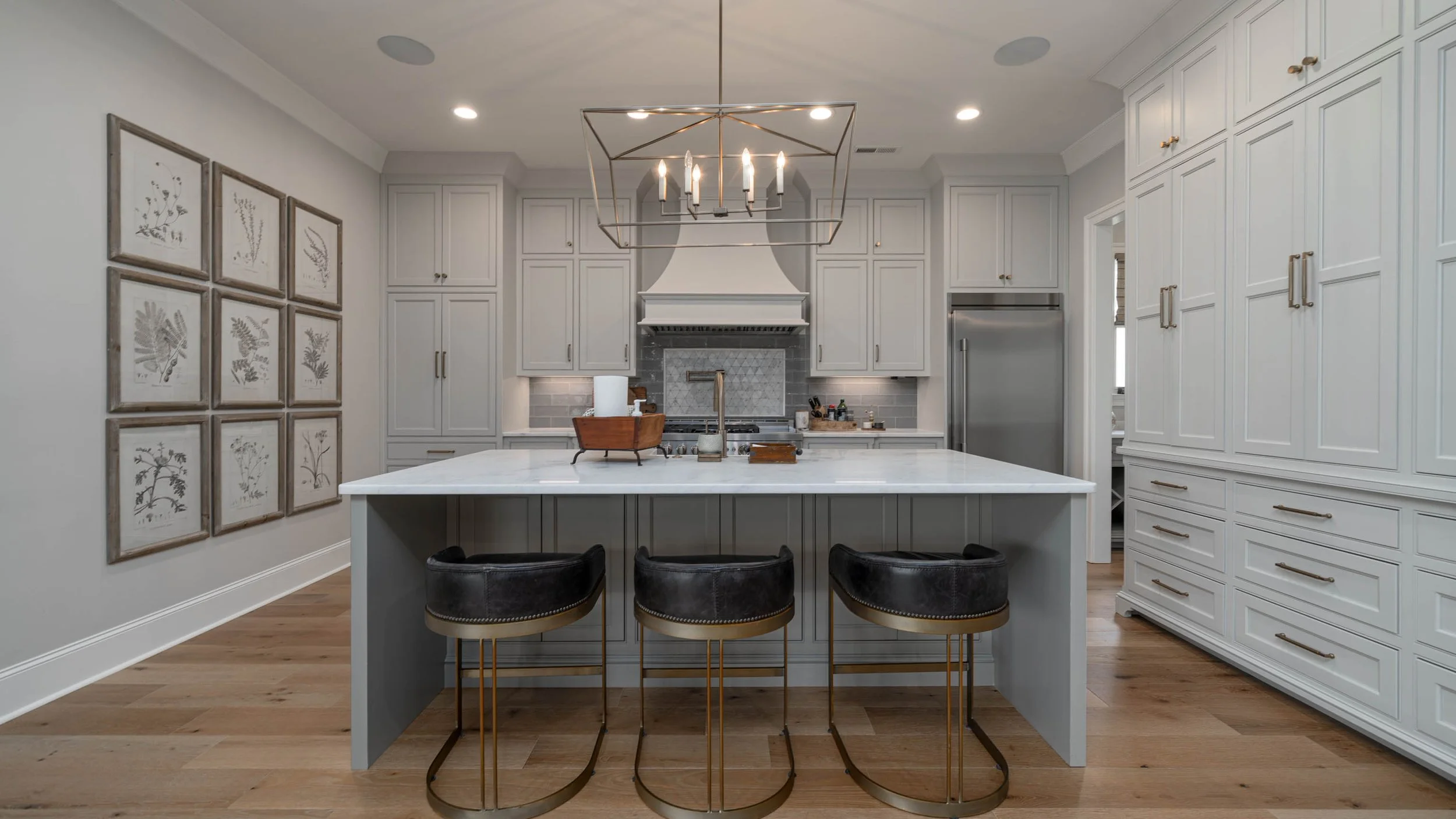 Modern kitchen with white cabinetry, a large marble island, black leather barstools with gold legs, wall art, and a chandelier above the island.