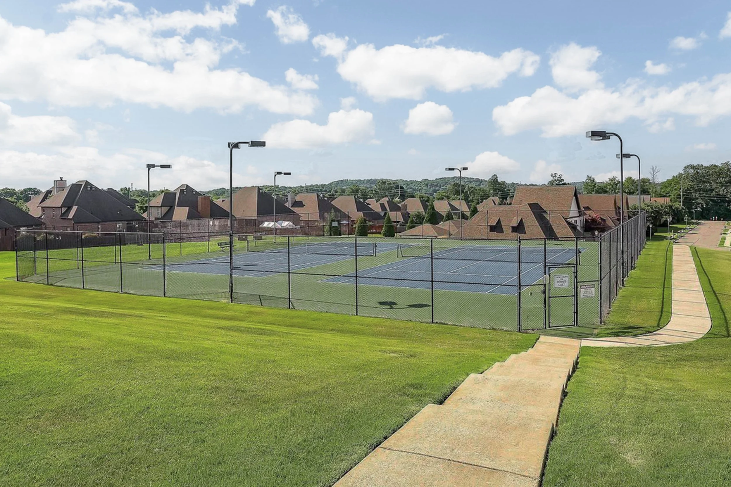 A fenced tennis court in a residential neighborhood with houses, grassy areas, and cloudy sky in the background.