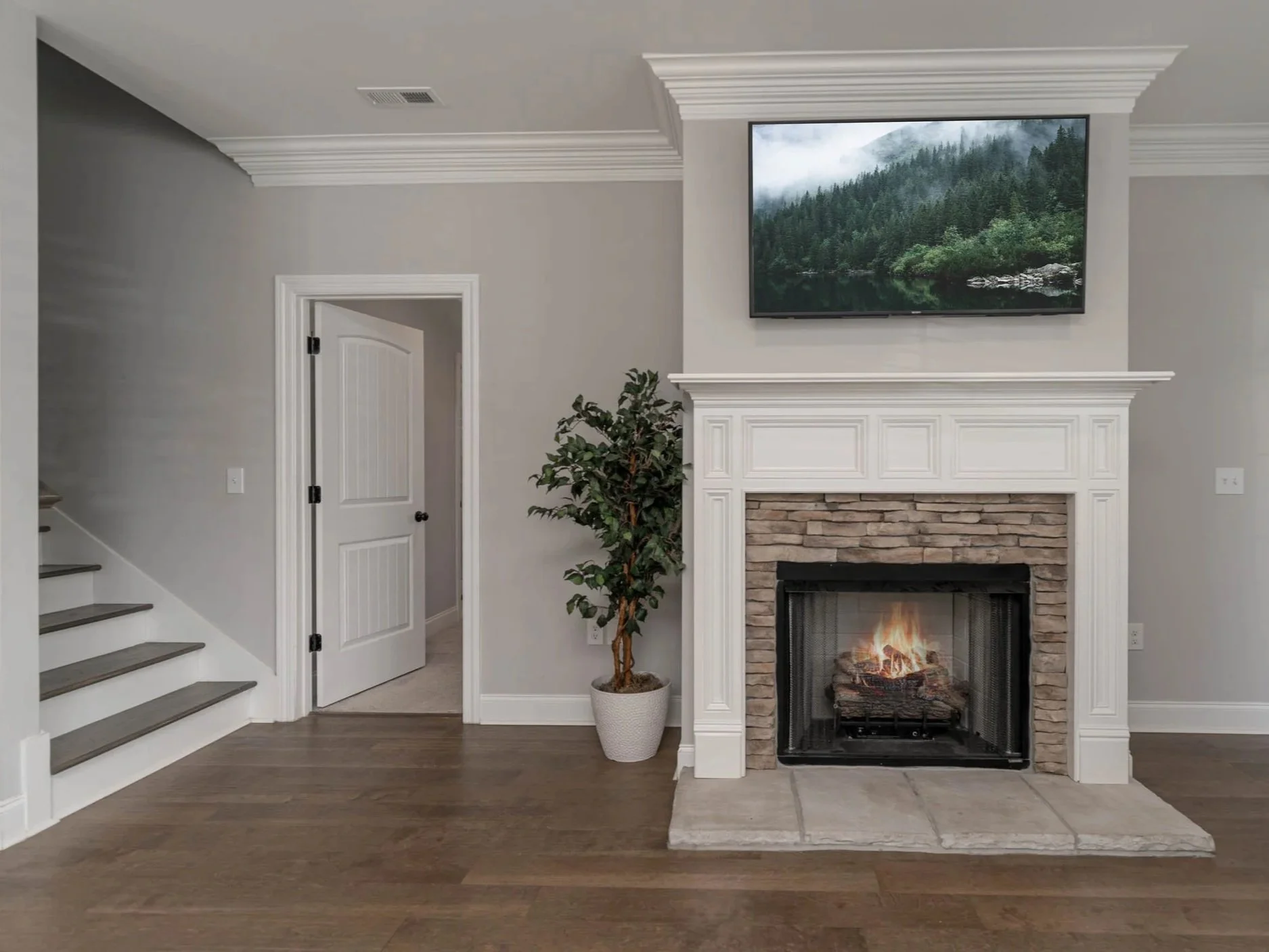 Living room with a white fireplace, a flat-screen TV above it displaying a landscape of a lake and forest, a potted plant to the side, and a staircase on the left.