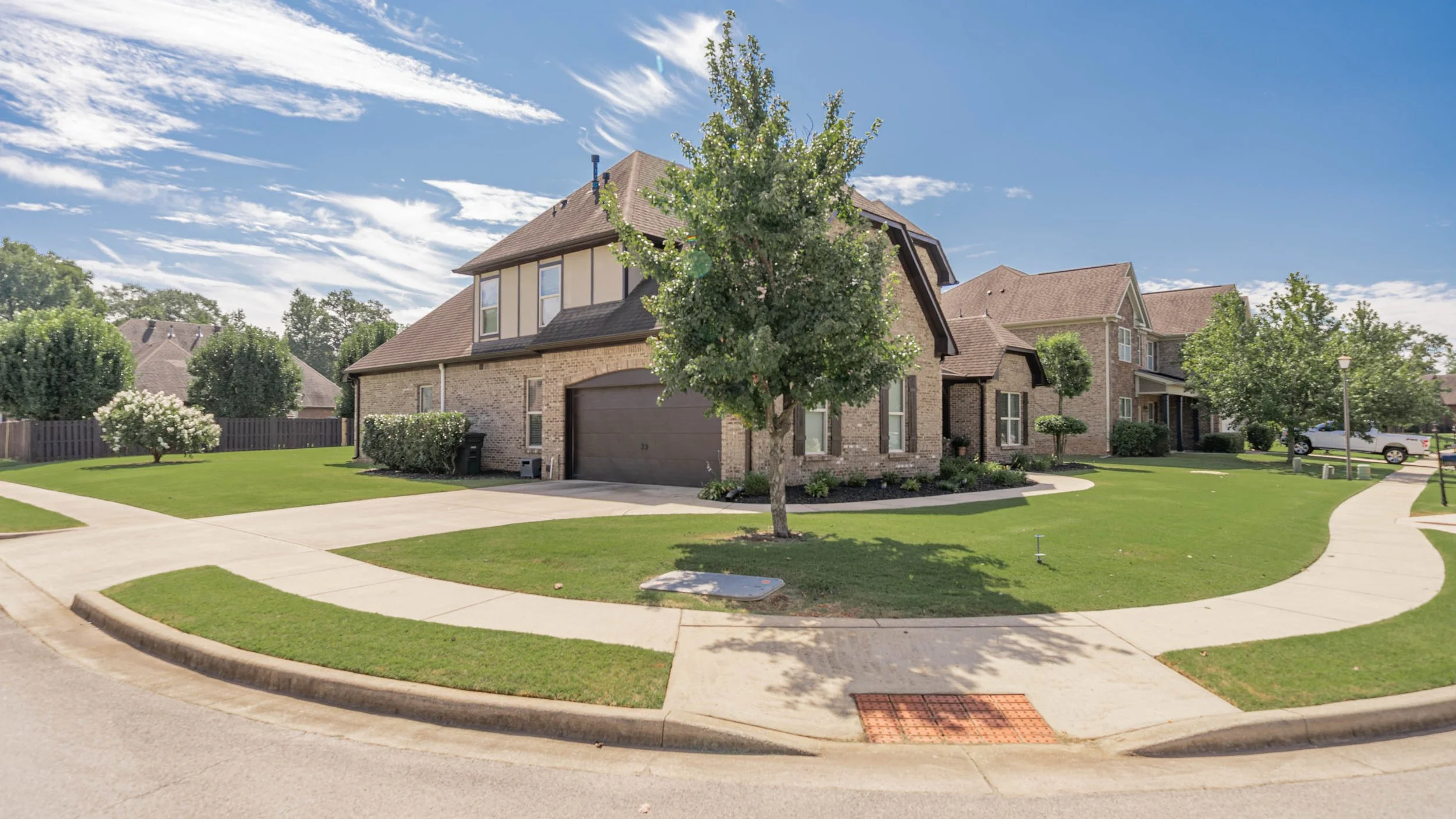 A suburban house with a brick facade, dark garage door, and front yard with trees and green lawn on a sunny day.