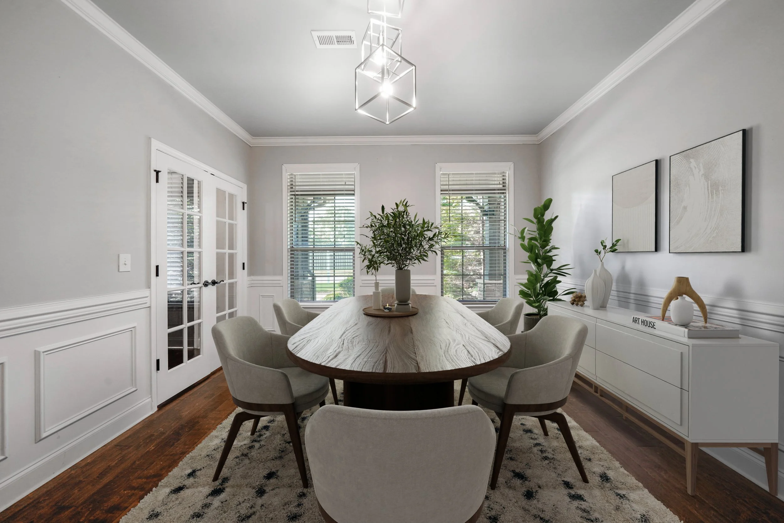 A modern dining room with a wooden table, beige chairs, a white sideboard, and minimal art on the wall. Three windows with blinds are behind the table, and a potted plant is in the corner.