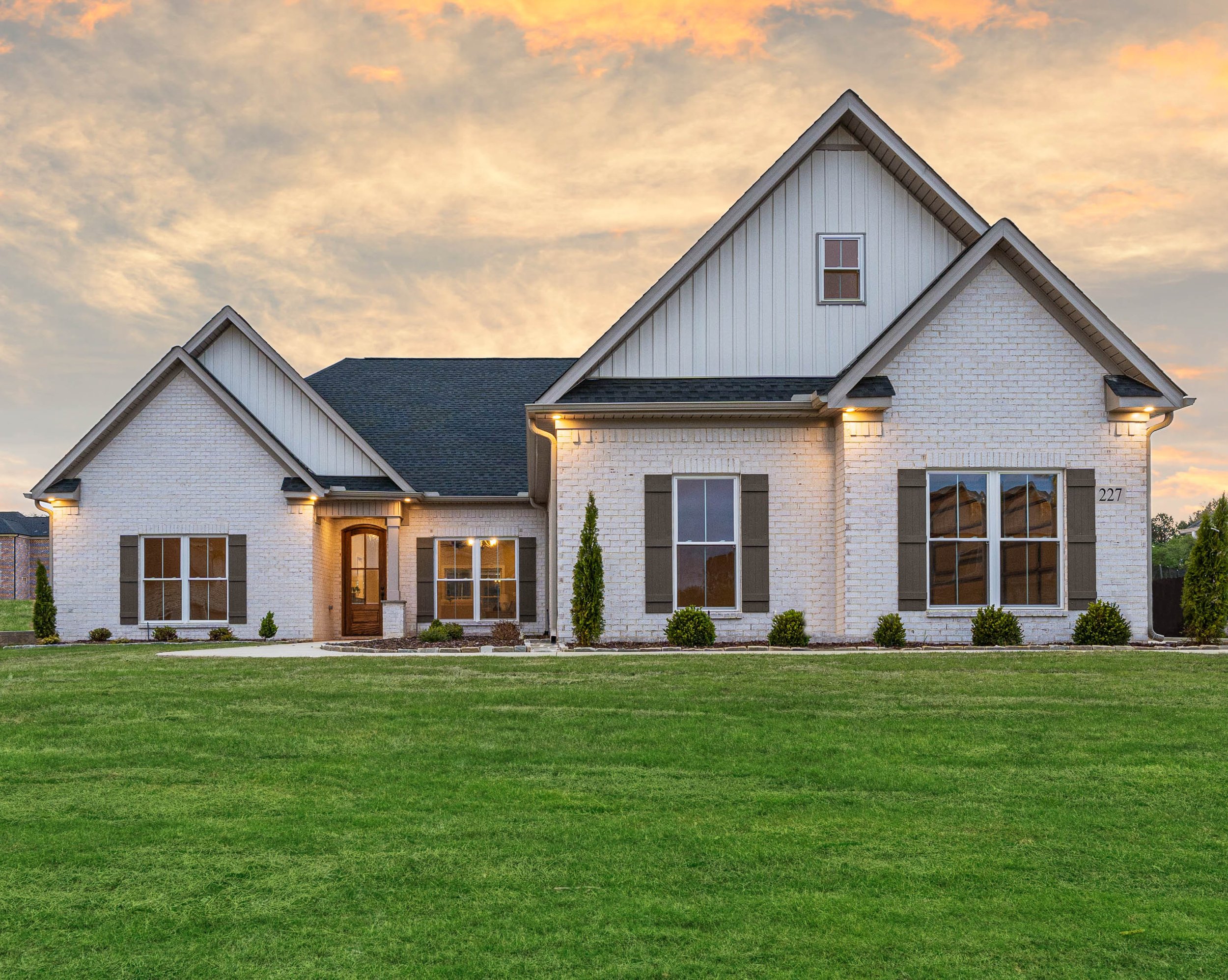 A modern two-story house with white brick exterior, large windows, and a front lawn at sunset.