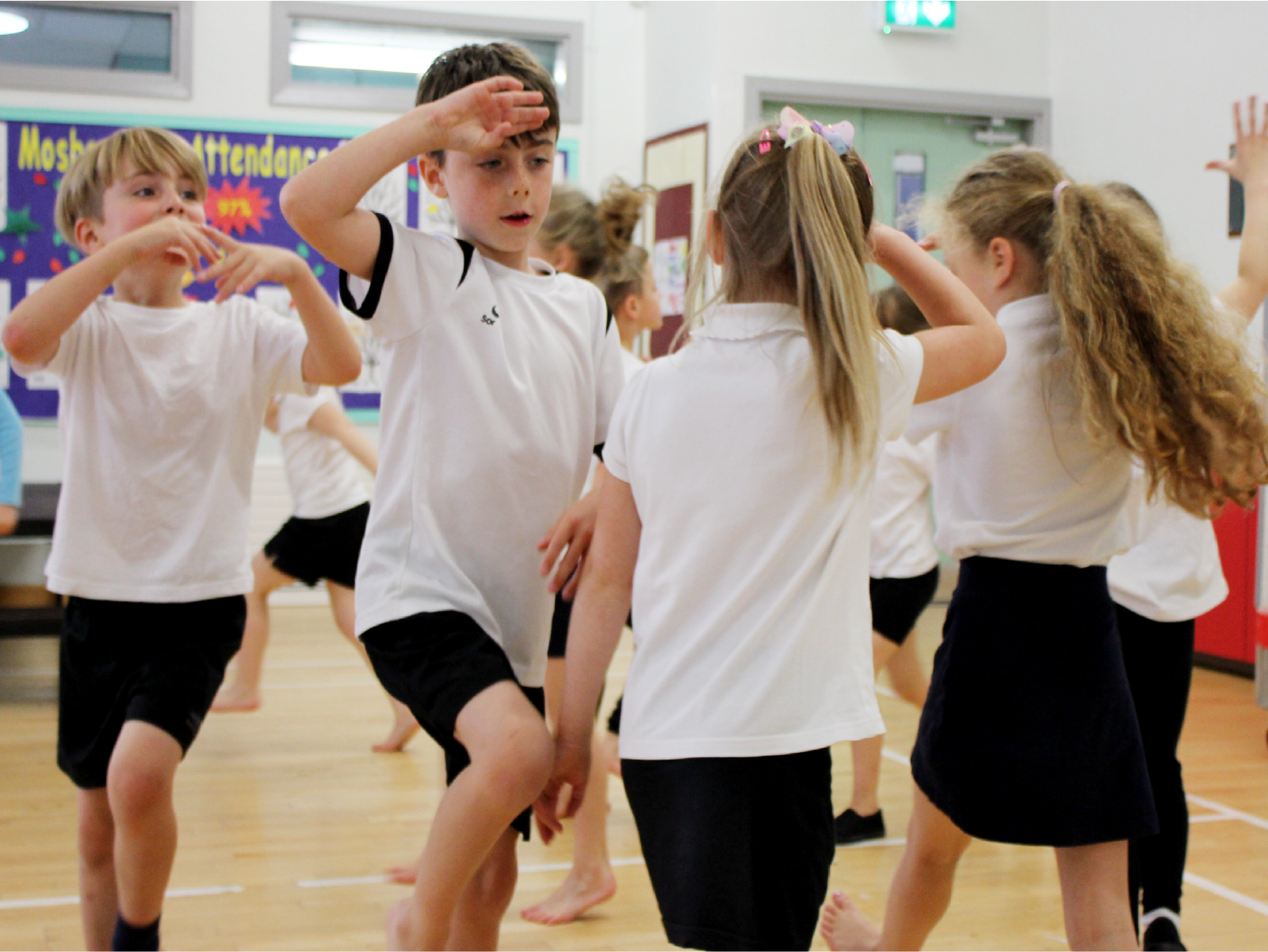 Children in a gymnasium participate in a dance or exercise class, wearing white shirts and black shorts, with some raising their hands and others moving energetically.