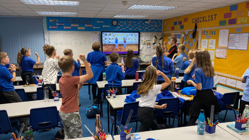 Elementary school classroom with students standing at desks, participating in a lesson while watching a dance video on a large TV screen. The room has educational posters, a whiteboard with math formulas, and a yellow bulletin board with a checklist chart.