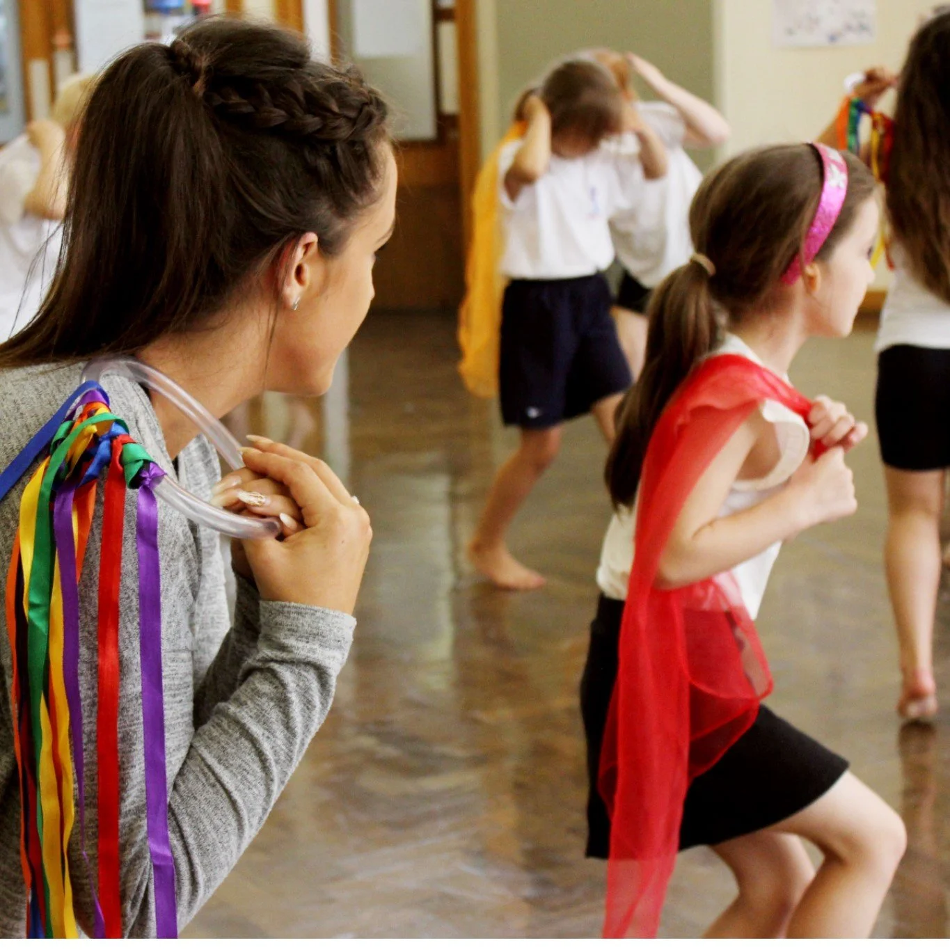 A woman with a rainbow-colored ribbon necklace watching children participate in an activity indoors.