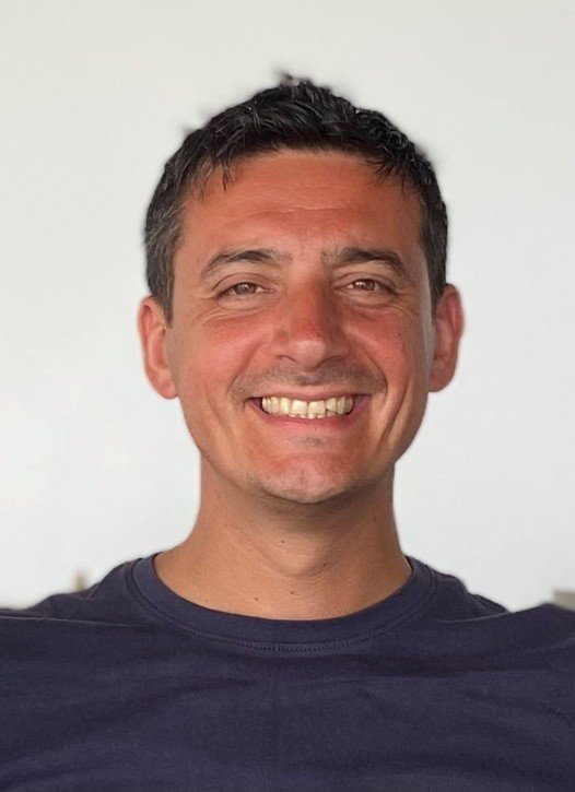 Smiling man with short dark hair, wearing a black shirt, against a plain white background.
