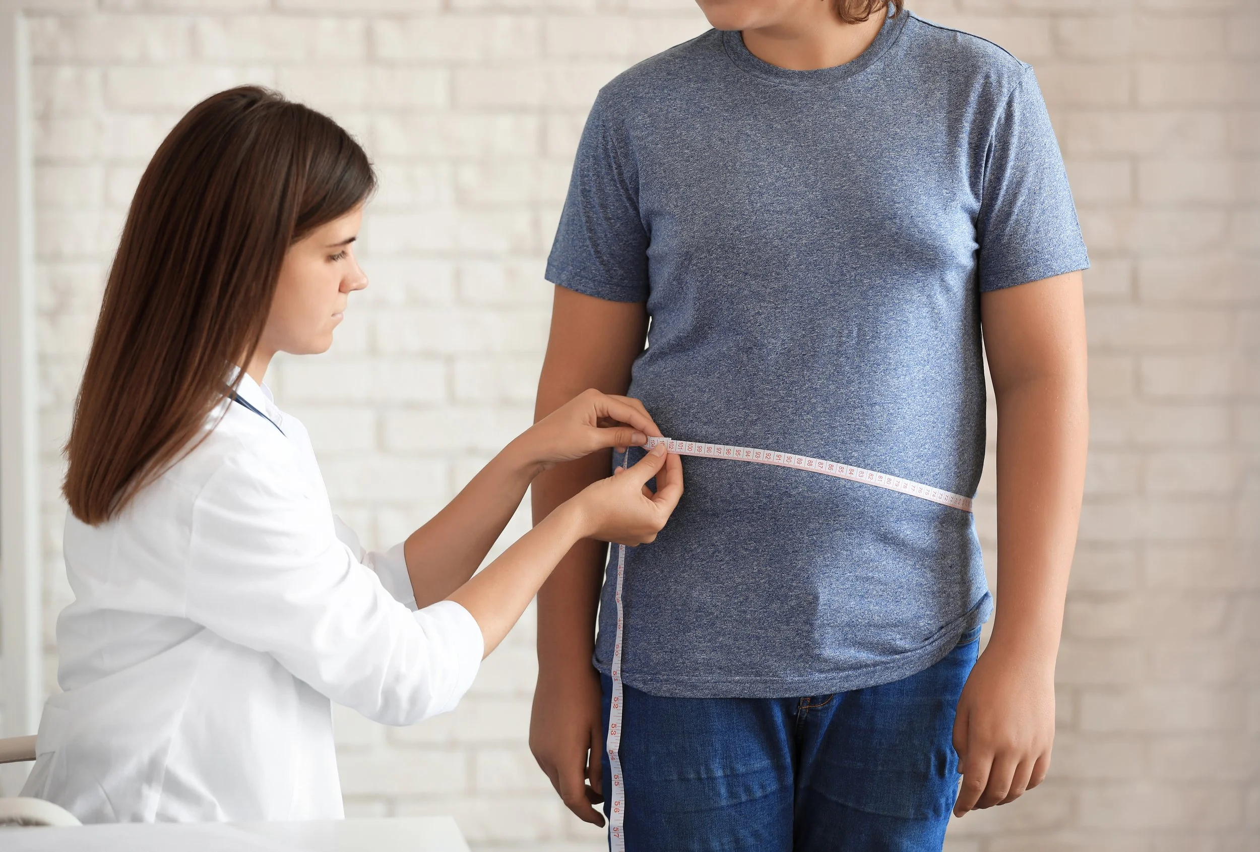A healthcare professional measures a young man's waist with a measuring tape as he stands in a room with a white brick wall.