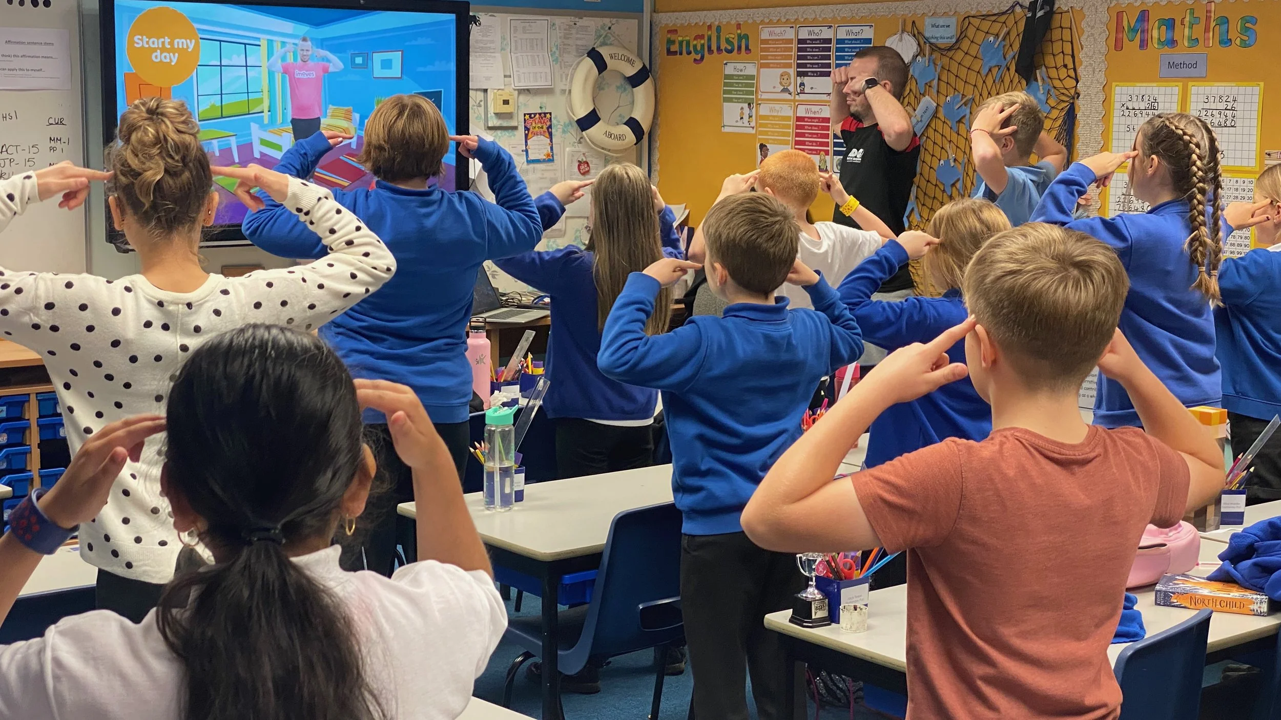 A classroom filled with students, mostly wearing blue uniforms, raising their right hands to their foreheads in a salute while a teacher stands near the whiteboard and a large screen displays a colorful scene. The classroom has educational posters and decorations on the wall.