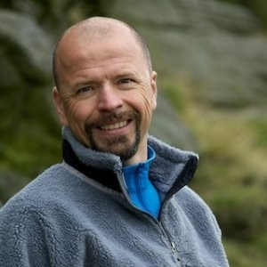A smiling man with a beard and short hair, wearing a blue jacket and gray fleece, outdoors with a rocky background.