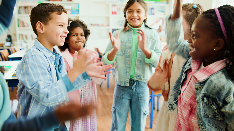 Children in a classroom clapping and smiling during a group activity.