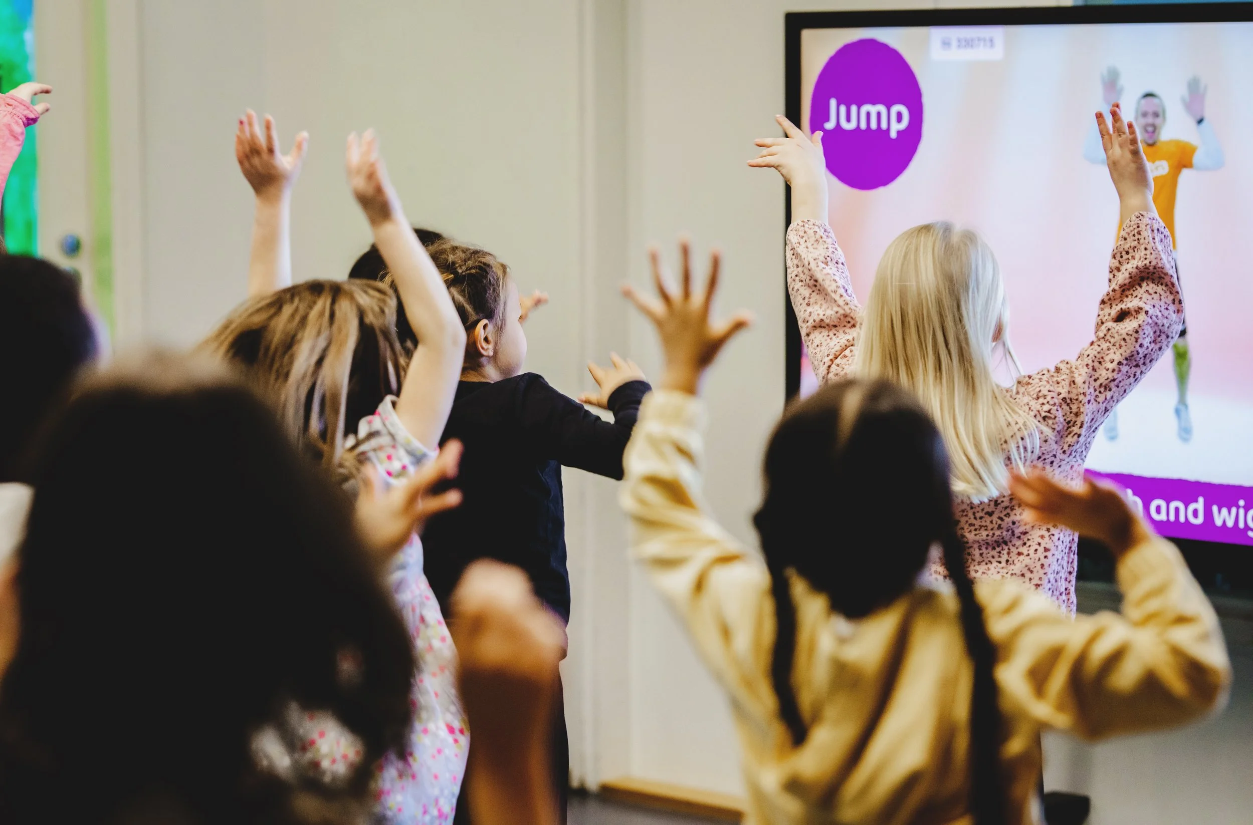 Children raising their hands in front of a large screen displaying a person in an orange shirt with an app icon labeled 'Jump'.