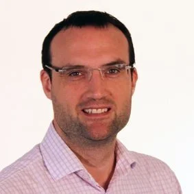 A man with glasses and receding hairline wearing a dark blazer and light shirt, smiling against a plain white background.