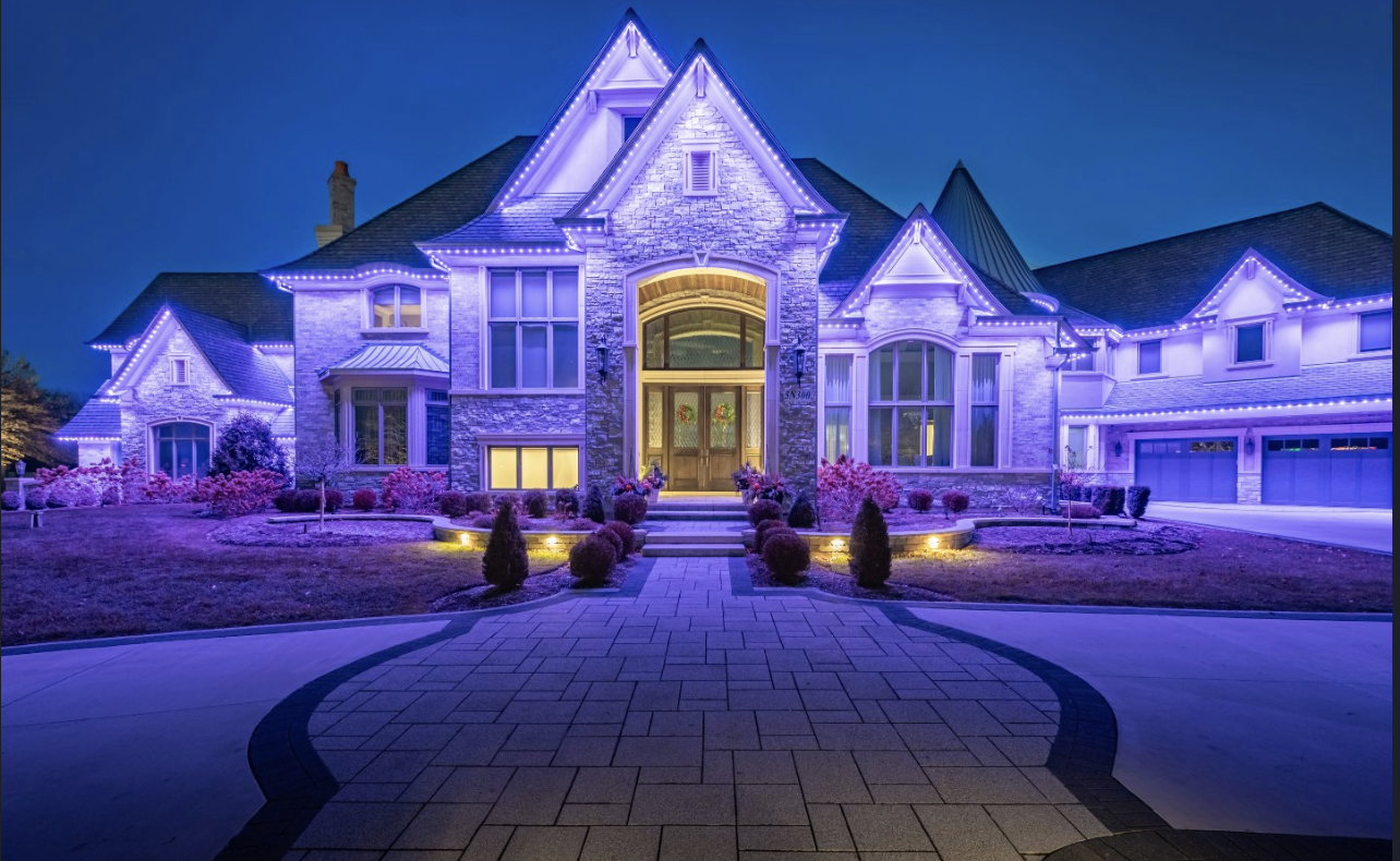 Large house illuminated with purple and white Christmas lights at night, with a landscaped front yard and brick walkway.