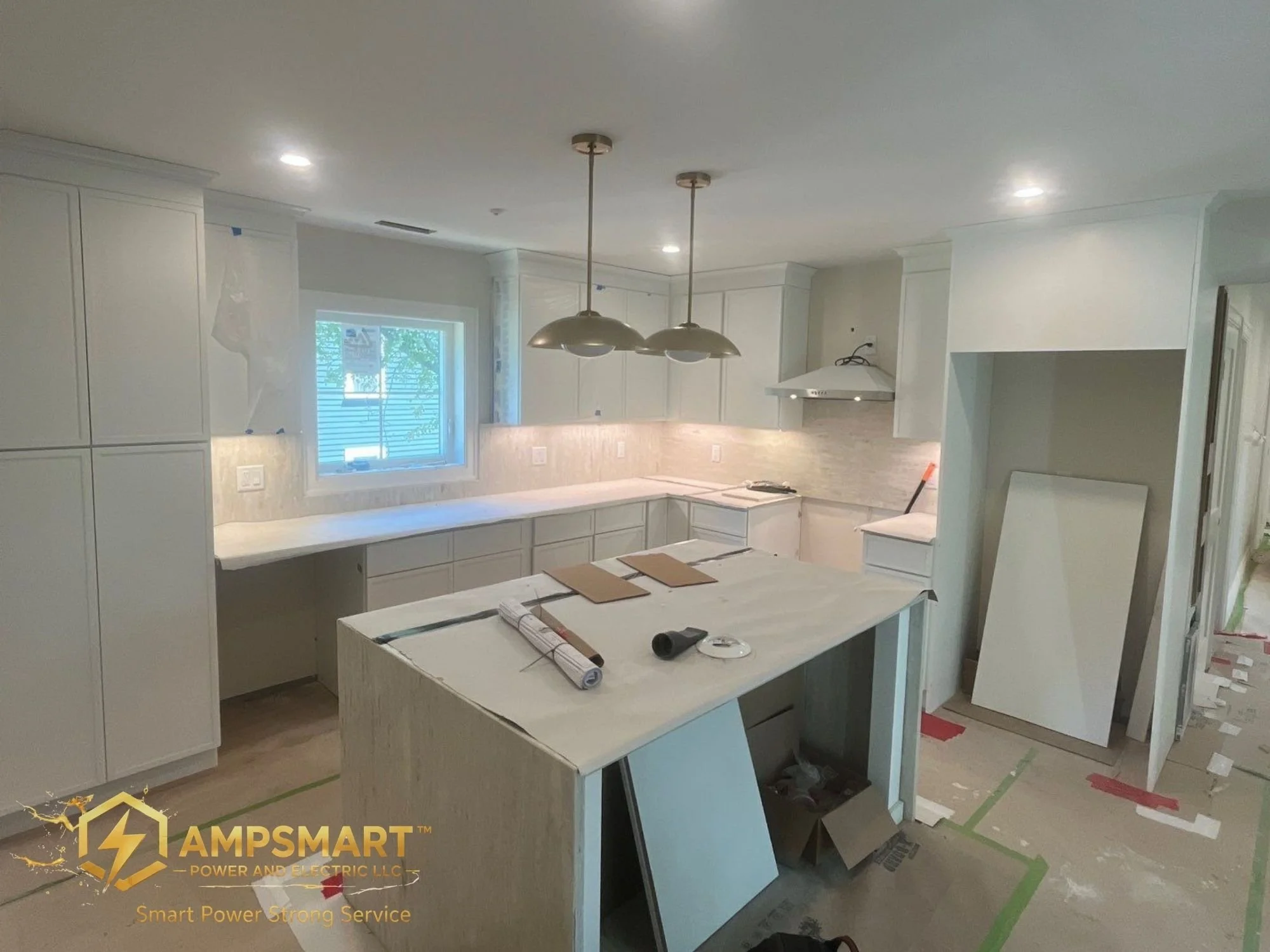 Kitchen under renovation with white cabinetry, a window, and two pendant lights hanging from the ceiling. Construction materials and tools are on the counter, and protective paper and tape are on the floor and walls.