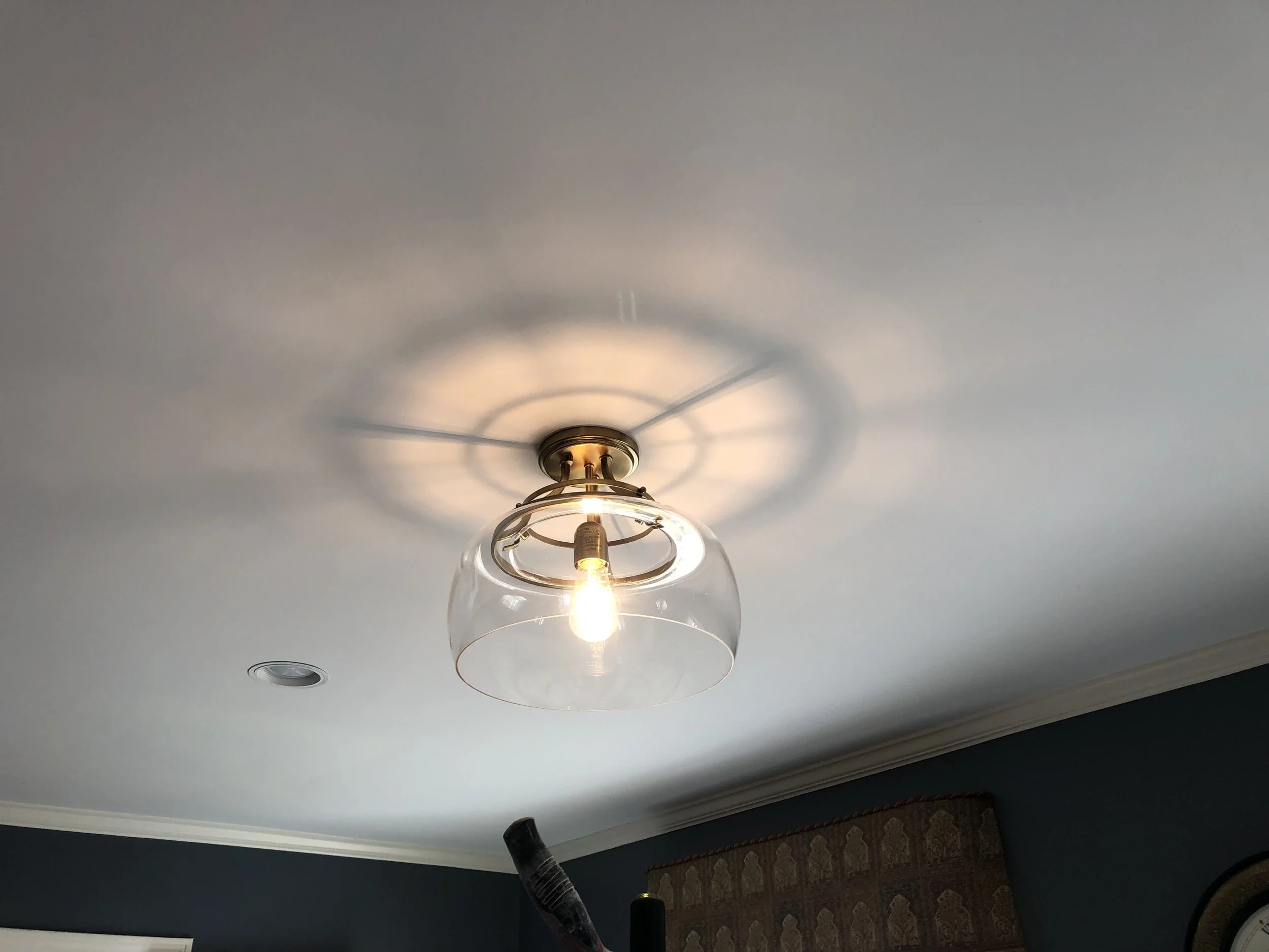 Ceiling light fixture with a glass shade and exposed light bulb, casting shadows on the ceiling, in a room with dark blue walls and decorative curtain.