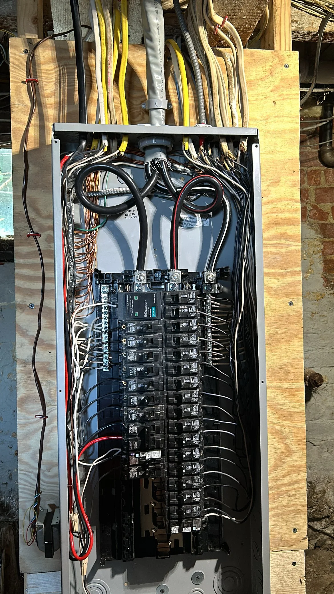 Open electrical panel with multiple circuit breakers and wiring inside, mounted on a wooden board.