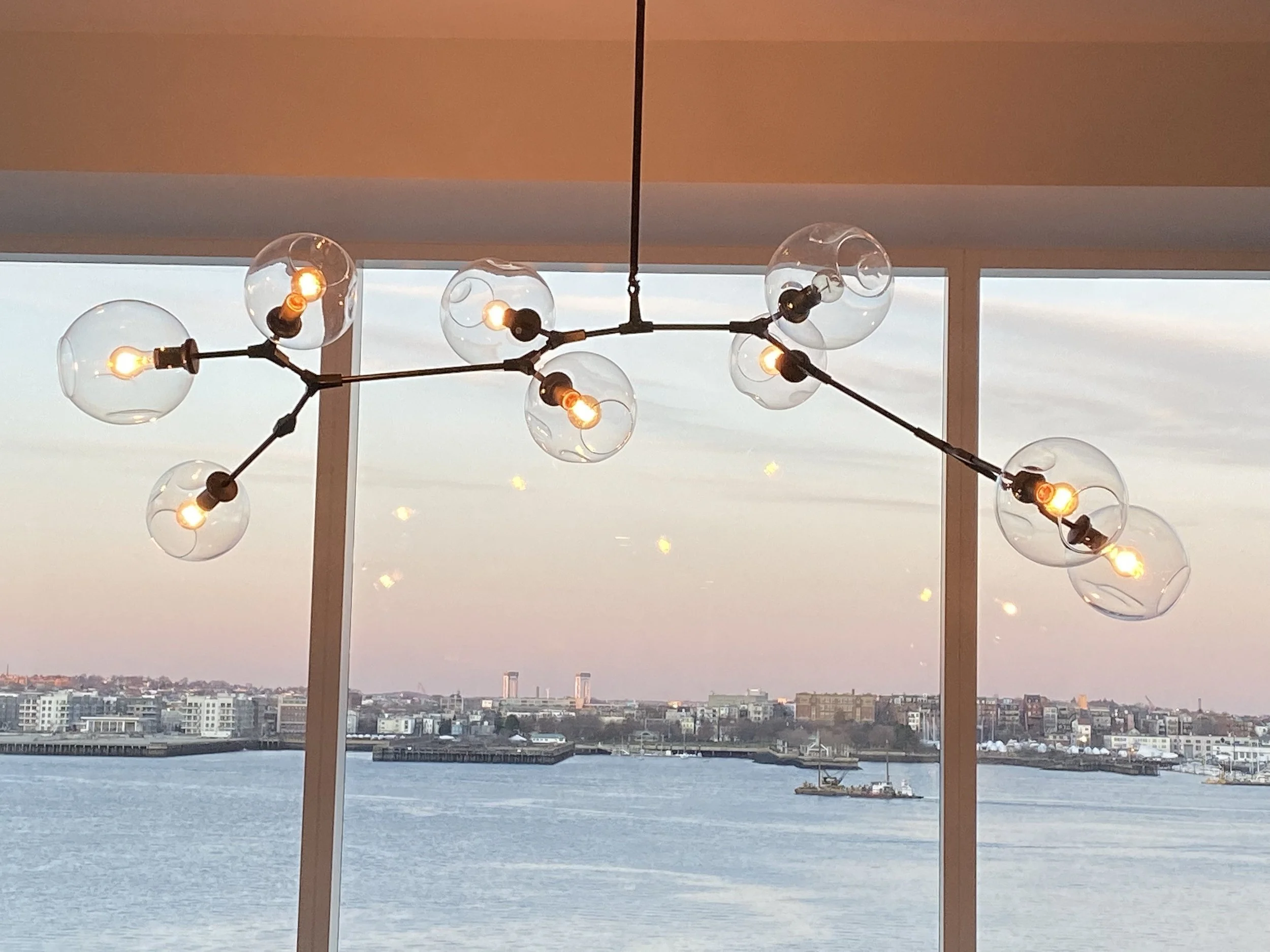 Modern chandelier with exposed bulbs hanging in front of a window overlooking a city river and skyline at sunset.