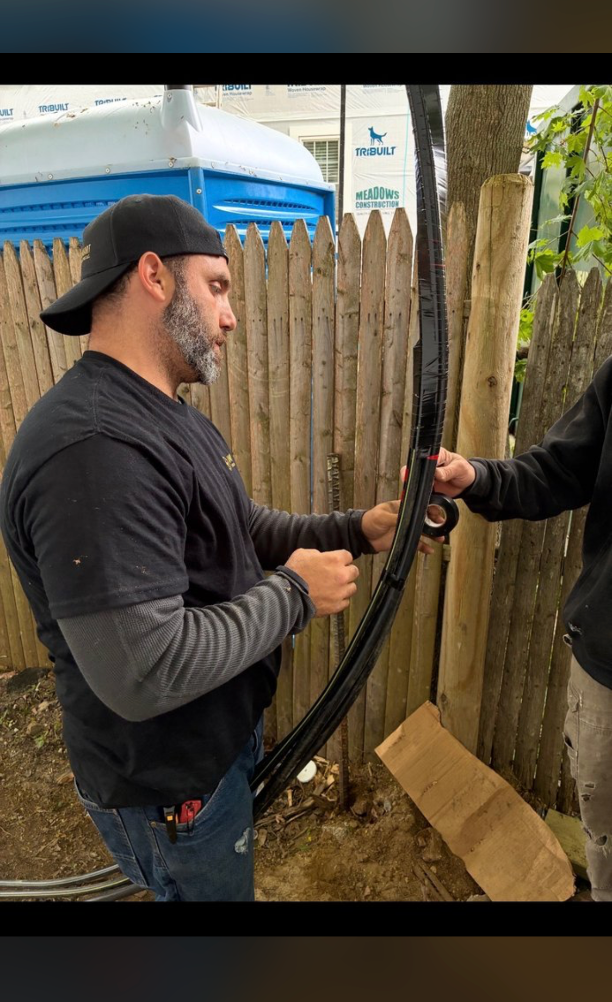 A man with a beard and a black cap working with equipment outdoors, standing near a wooden fence. He is wearing a black t-shirt over a gray long sleeve shirt and blue jeans.