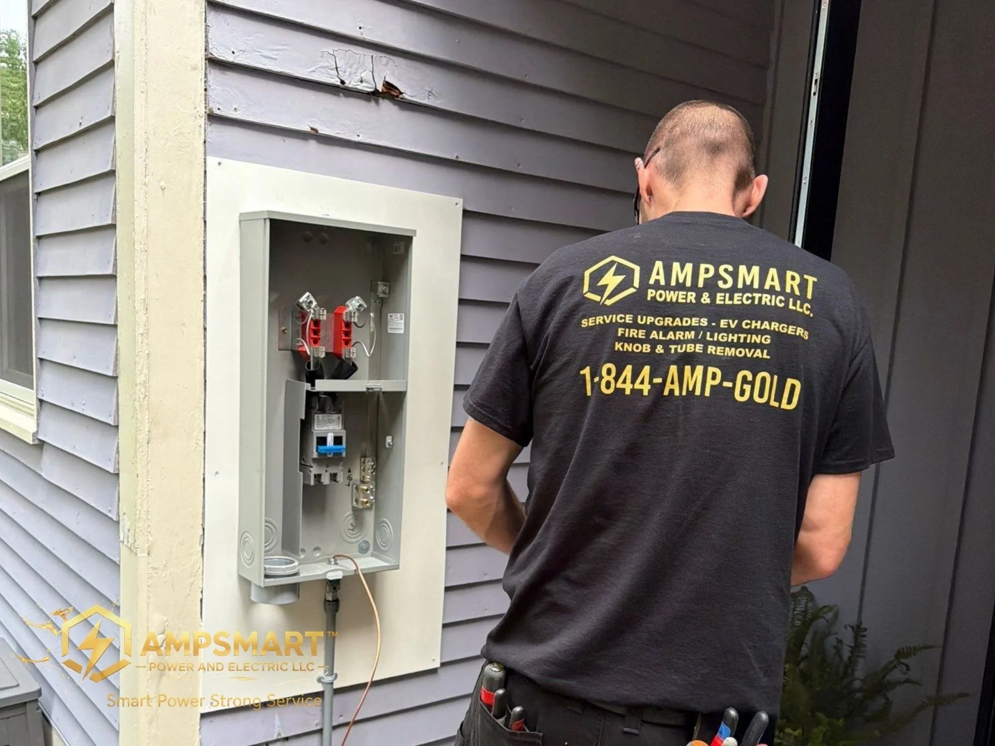 A technician with a black shirt that reads "AMPSMART POWER & ELECTRIC LLC" is working on an outdoor electrical box attached to the side of a house with gray siding. The electrical box is open, showing wiring inside.