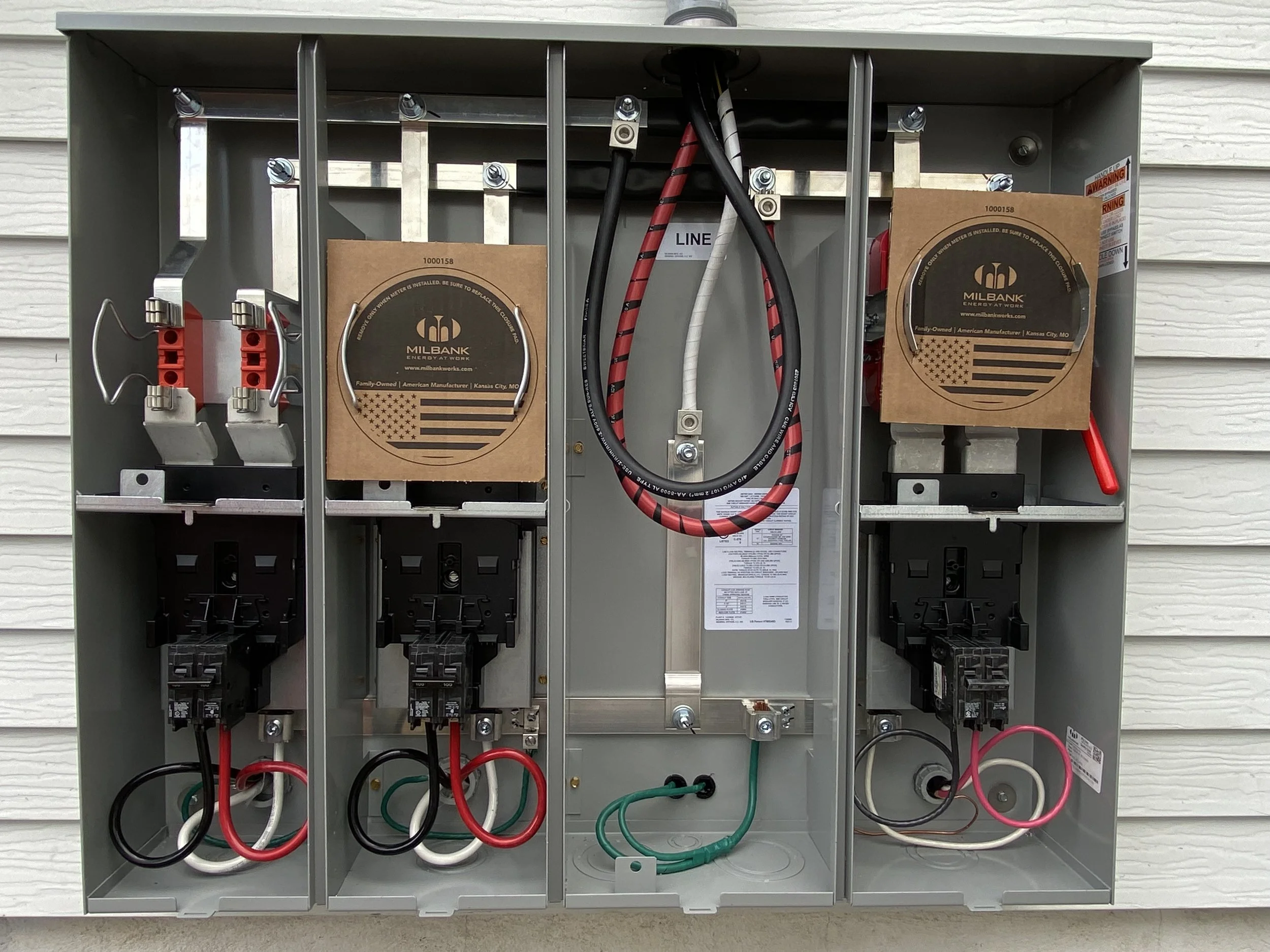 Electrical breaker box with wiring and components mounted inside, set against a white siding wall.