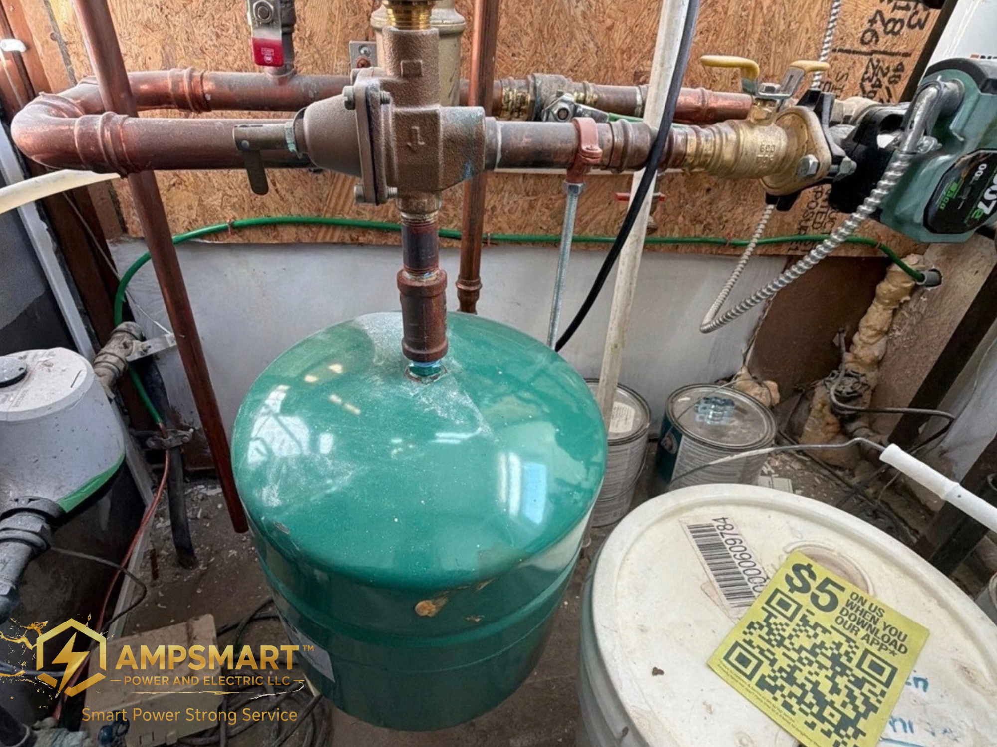 A manifold with copper pipes connected to it, situated in a construction or utility room with a wooden wall in the background. There are tools, cans, and a QR code on a yellow card nearby.