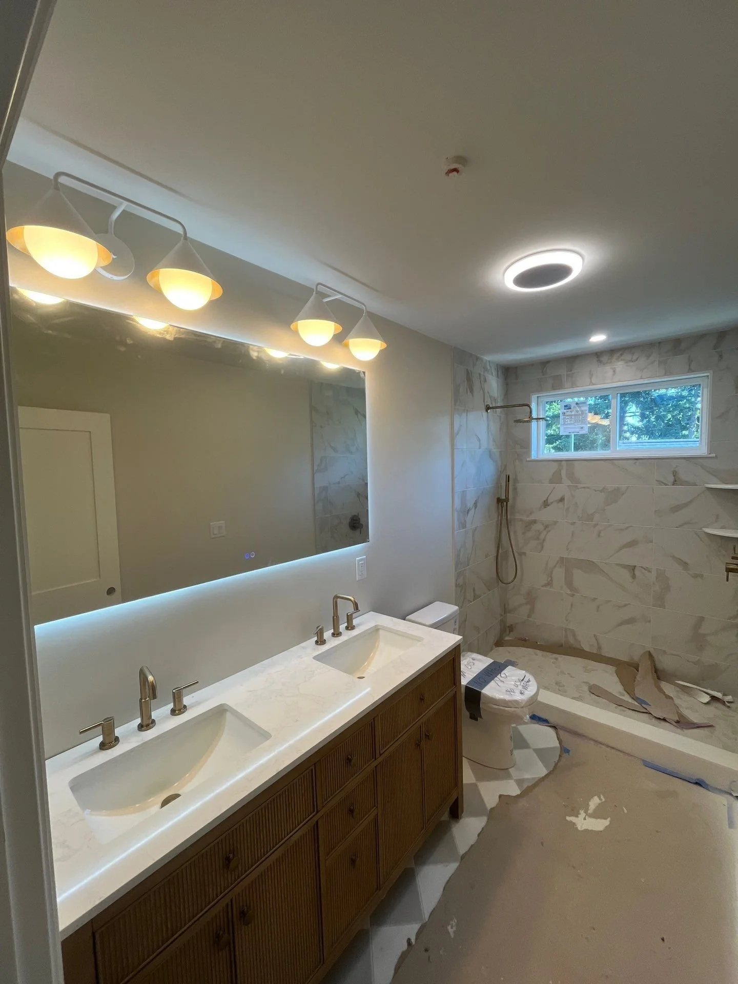 Bathroom under renovation with double sink vanity, large mirror, wall-mounted lights, a toilet covered in protective wrapping, a shower area with a window, and construction debris on the floor.
