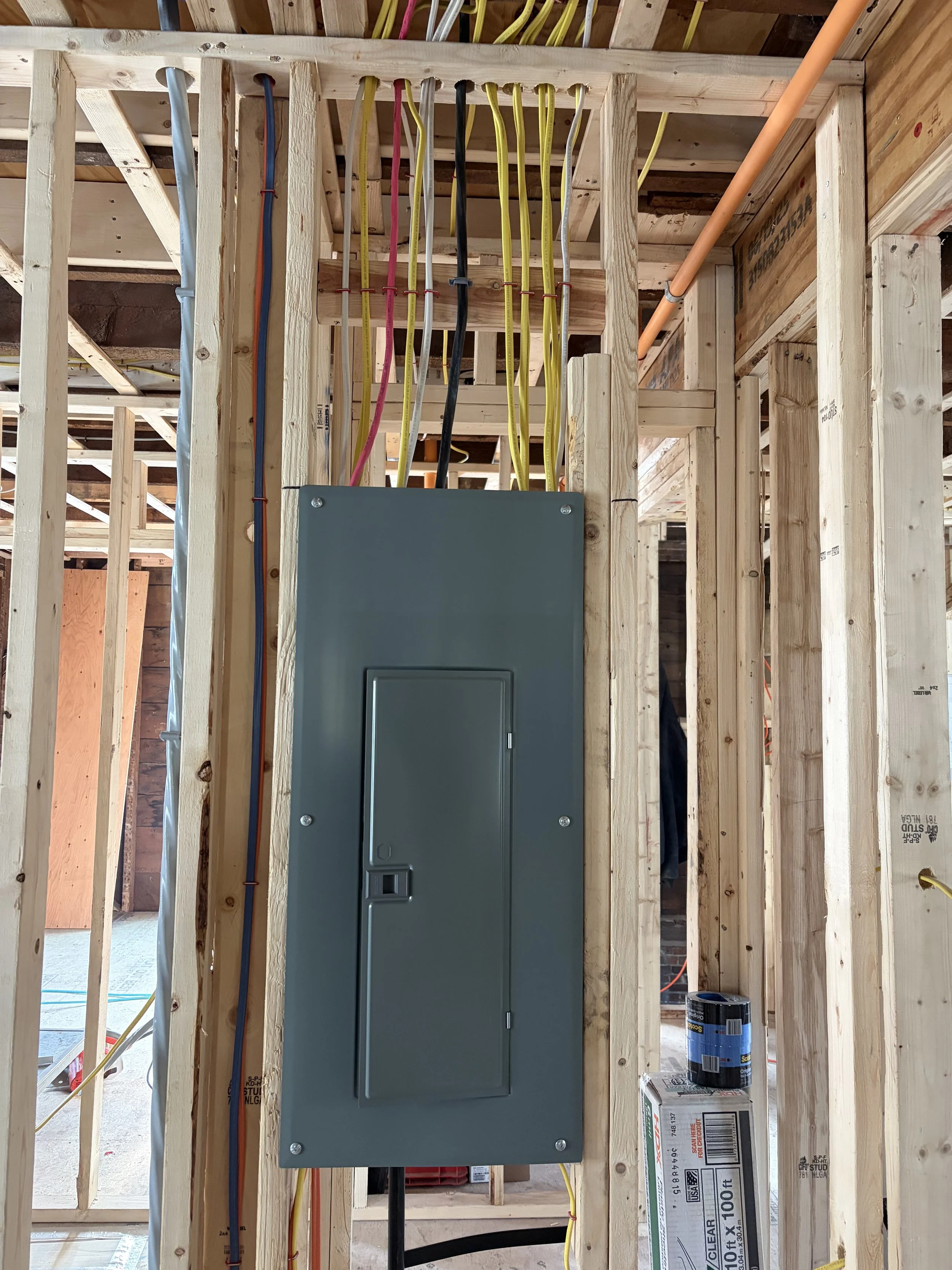 Electrical panel installed in the construction framing with color-coded wiring running to the ceiling for home wiring system.