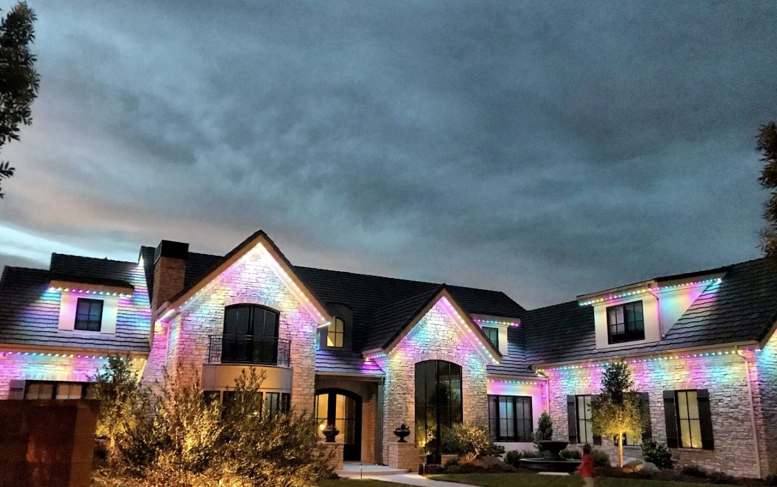 A large house decorated with colorful Christmas or holiday lights on the roofline and around windows, with a dark, cloudy sky in the background.
