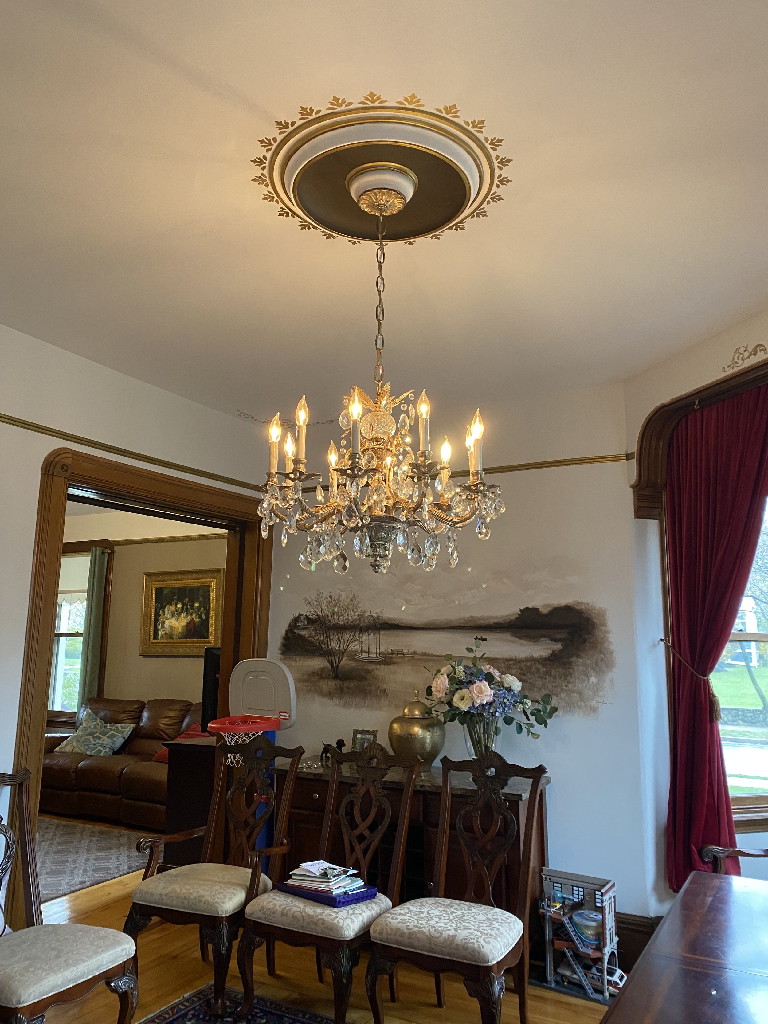 A chandelier hanging from ornate ceiling medallion in a dining room, with a large floral arrangement, a wall painting, and a small basketball hoop in the background.
