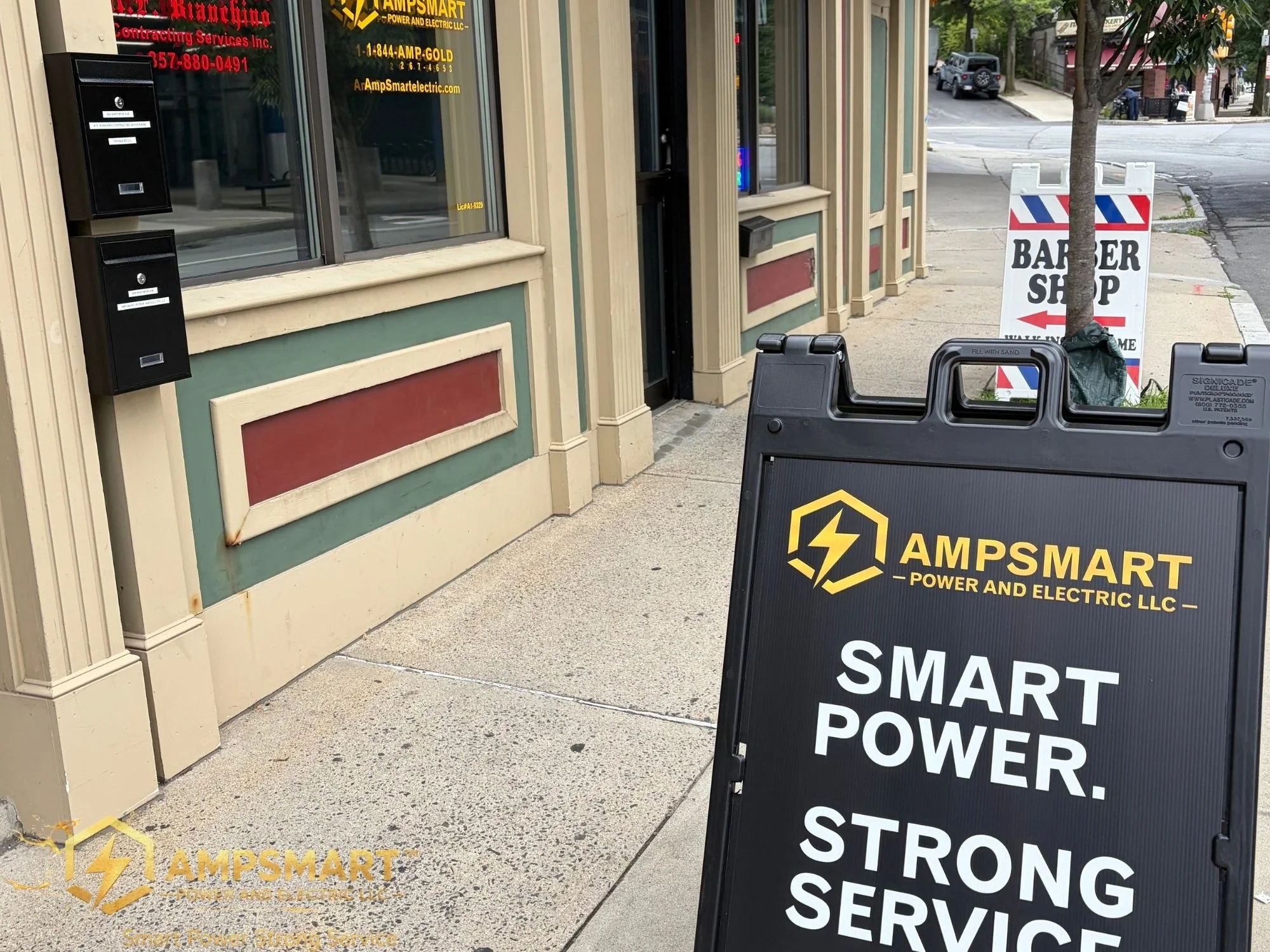A sidewalk scene outside a storefront with a window and a door, advertising electrical services by AmpSmart. There is a sign on the sidewalk that reads 'Barber Shop' with an arrow pointing left. A tree is also present near the sidewalk.