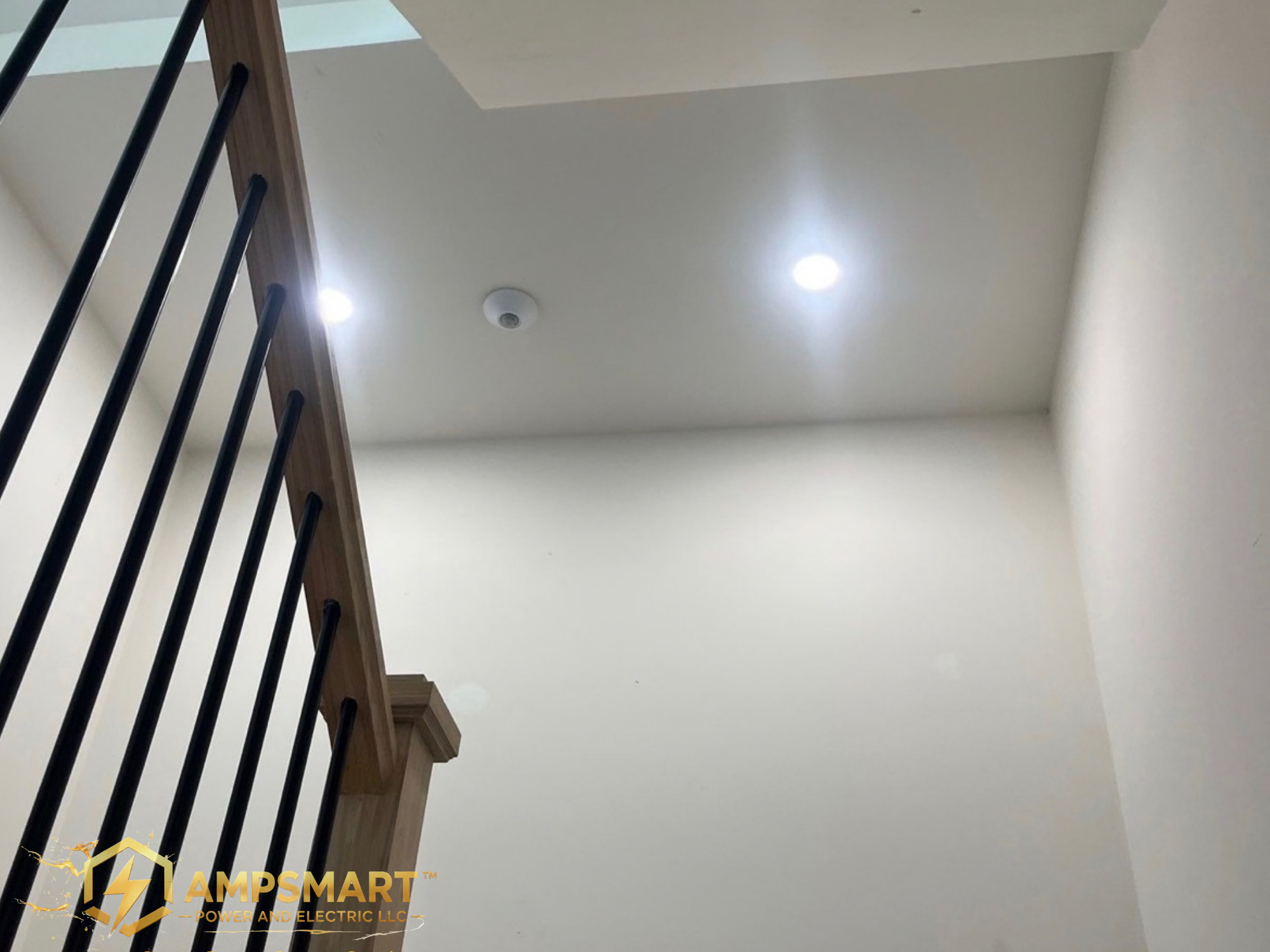 Interior view of a staircase with black metal balusters and a wooden handrail, upward-looking ceiling with recessed lighting and a smoke detector.