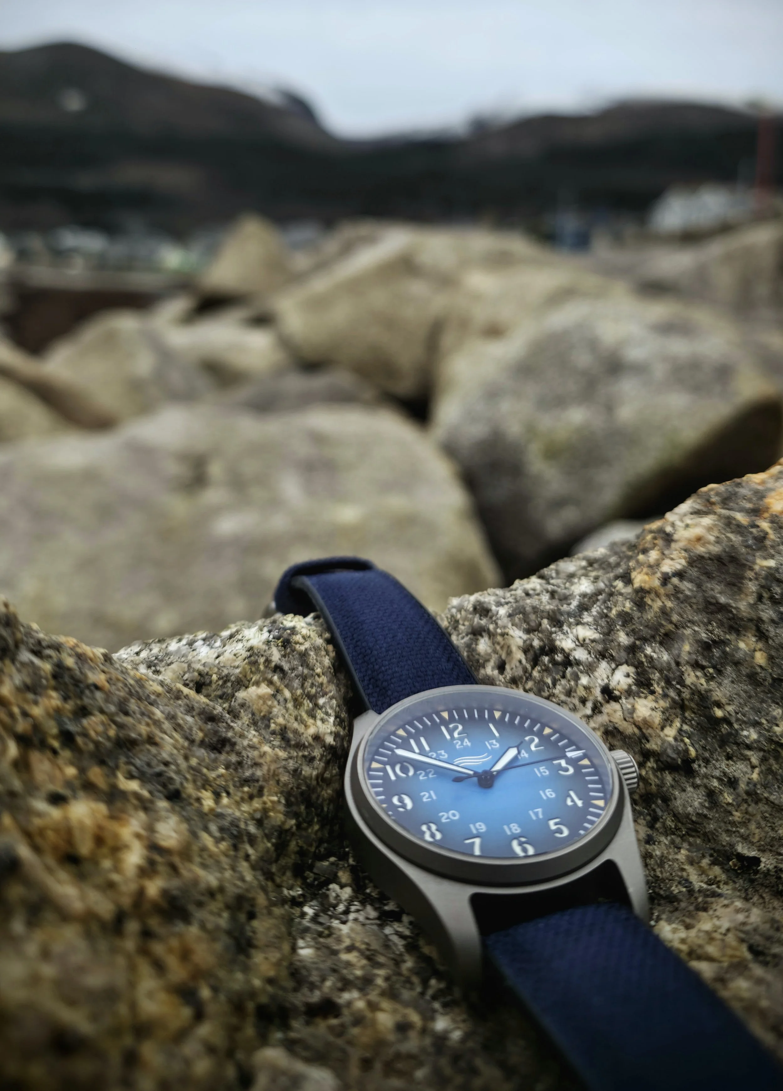 A wristwatch with a blue face and black strap resting on a rough, brown rock, with blurred rocks and distant hills in the background.