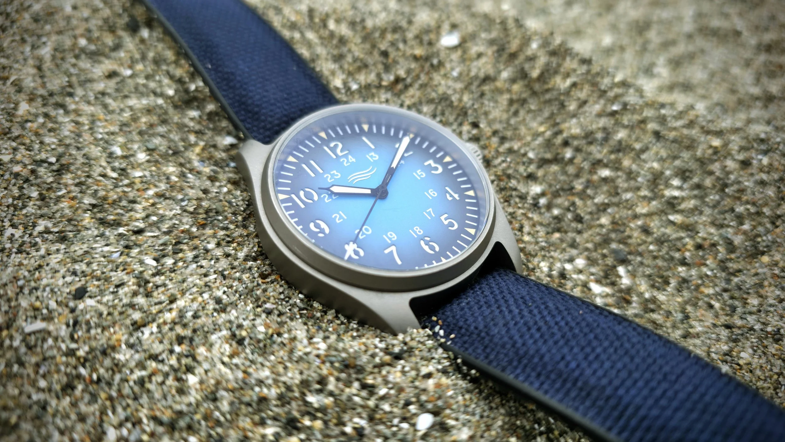 Wristwatch with a blue face and black strap lying on sand beach.