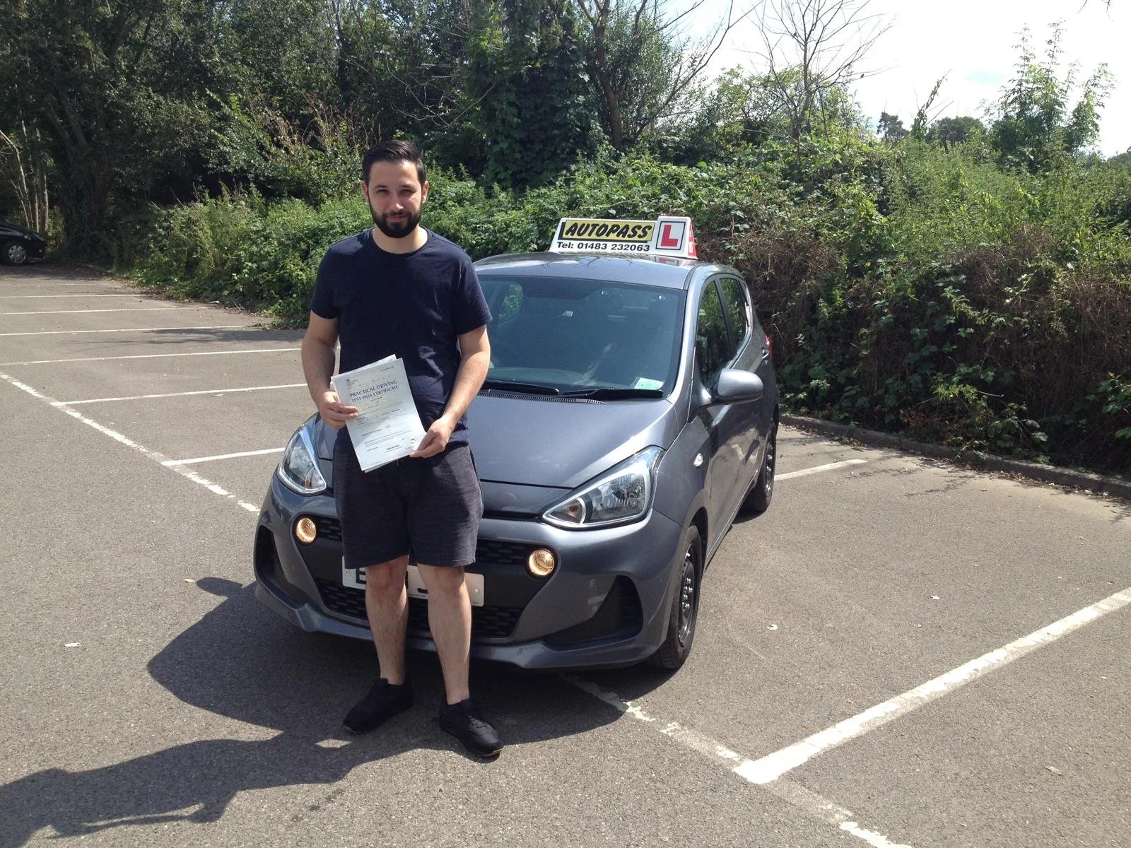 A man holding a driving license and a certificate standing in front of a silver hatchback car with an 'L' learner sign on the roof, parked in a parking lot surrounded by trees.