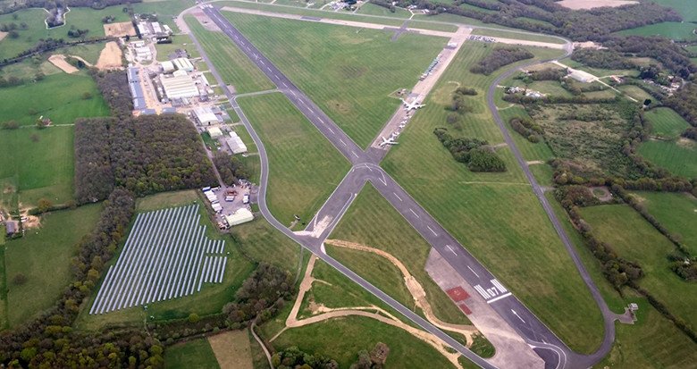 Aerial view of an airport runway with taxiways, surrounding green fields, and some buildings nearby.