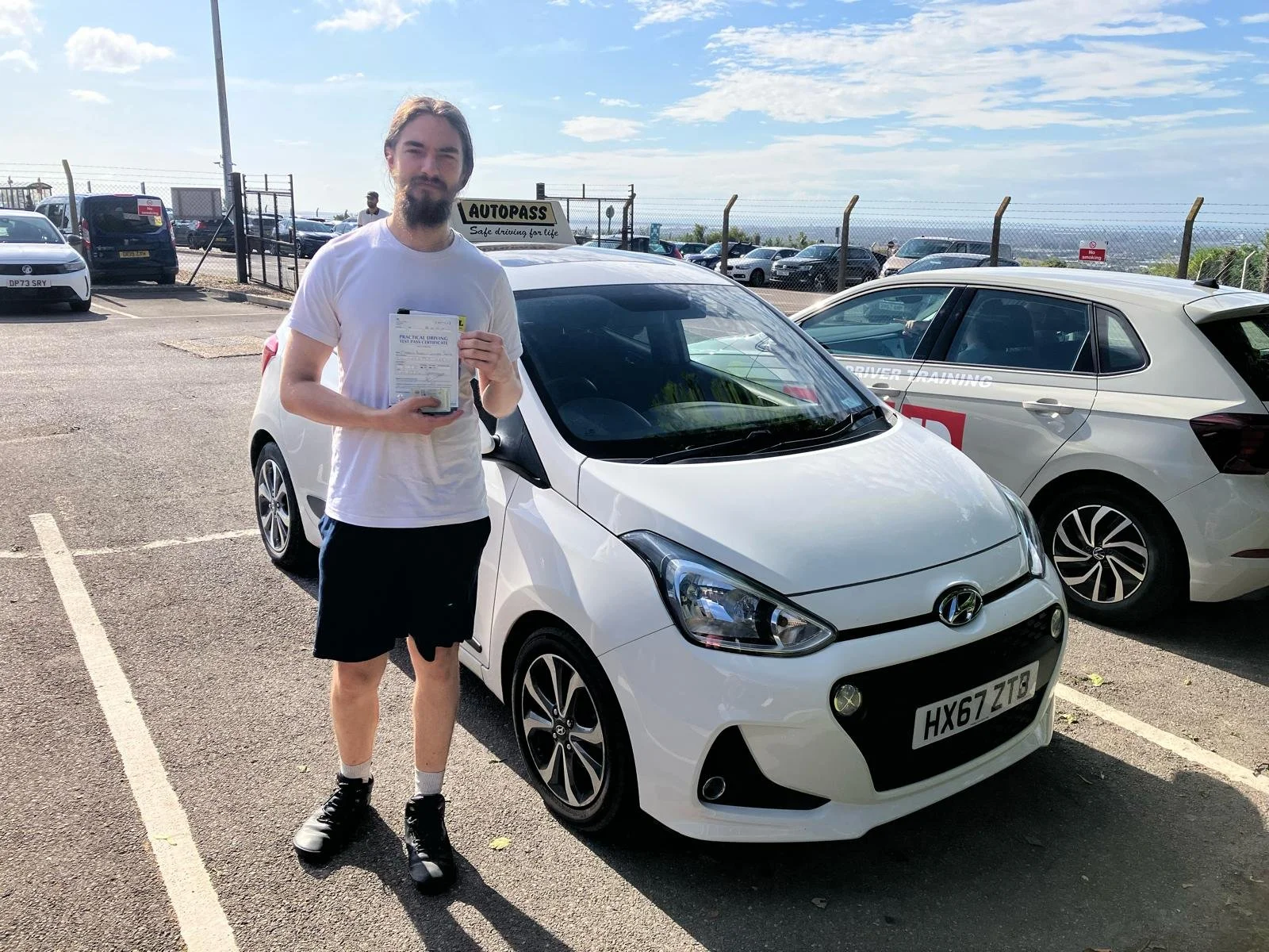 A man holding a driving test paper standing next to a white Hyundai i10 car in a parking lot, with a car behind labeled 'Driver Training' and a fence with parked cars in the background.