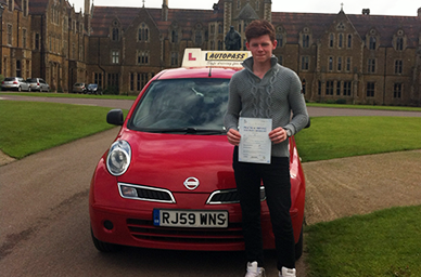 Young man standing next to a red learner driver car holding a certificate, with a historic building in the background.