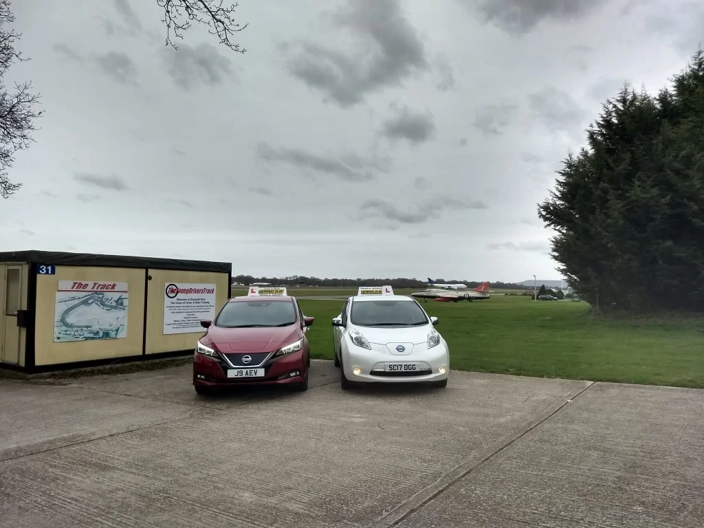 Two driving school cars, one maroon and one white, parked on a tarmac near a grass field with small planes in the background and a cloudy sky overhead.