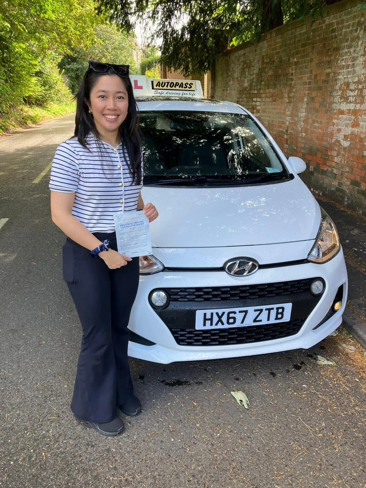 A woman standing next to a white Hyundai driving school car holding a certificate. The car has a sign on top indicating it's for driving lessons. The woman is smiling, wearing a striped shirt, and sunglasses on her head, with trees and a brick wall in the background.