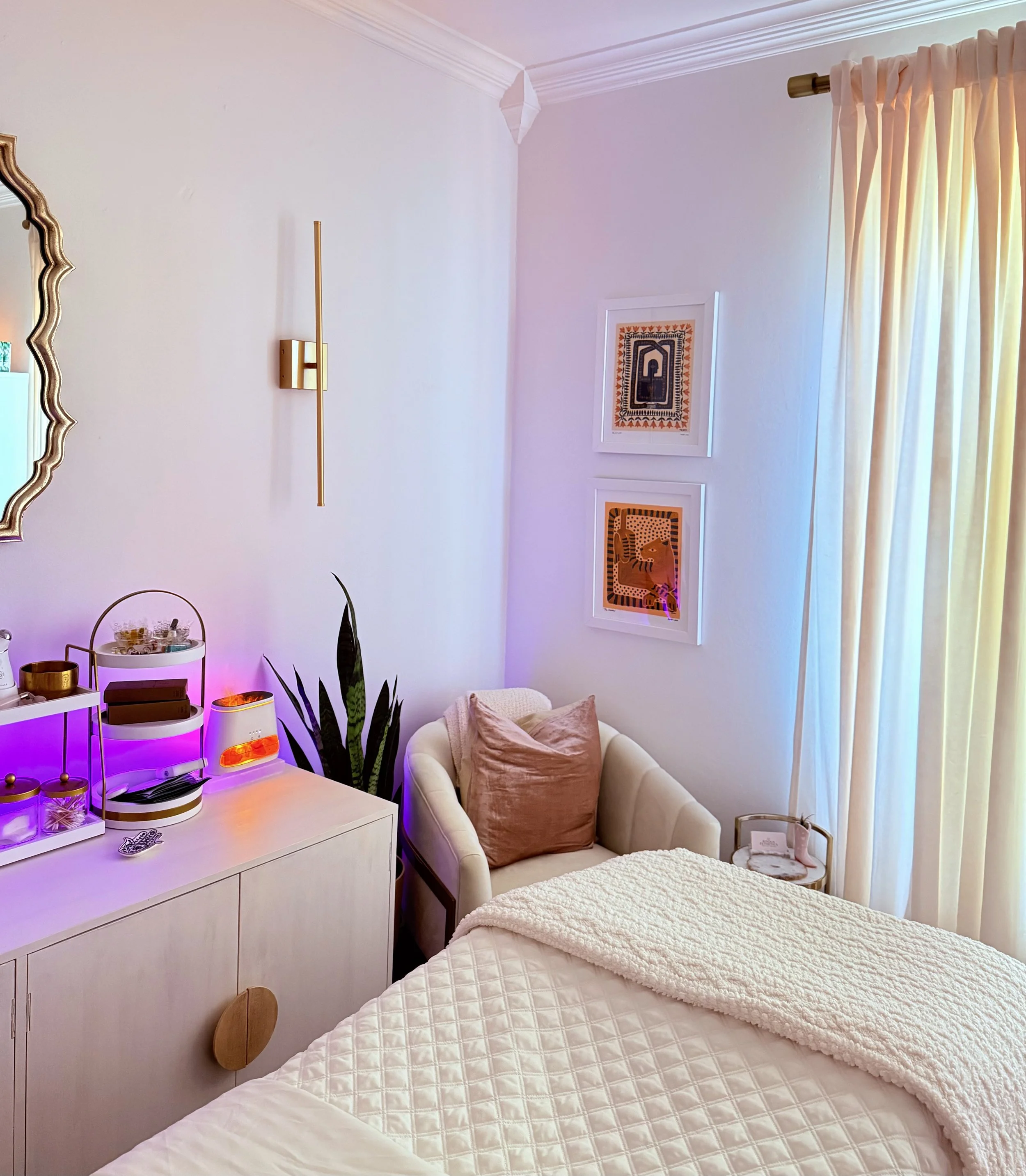 A cozy bedroom with two neatly made beds with white quilts, a cream armchair with a blush pillow, a white dresser with colored candles and skincare items, and framed artwork on the wall. There's a window with cream curtains and natural light, and houseplants near the dresser.