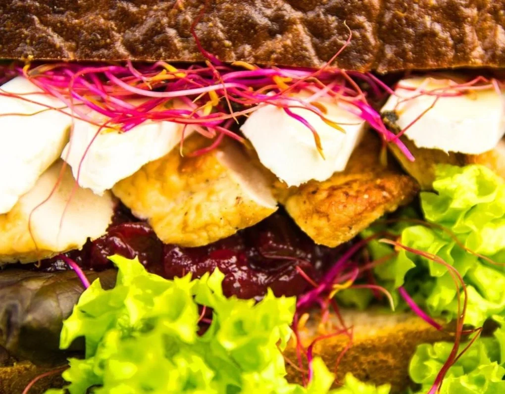 Close-up of a sandwich with fried chicken, leafy greens, sliced cheese, and colorful sprouts inside whole grain bread.
