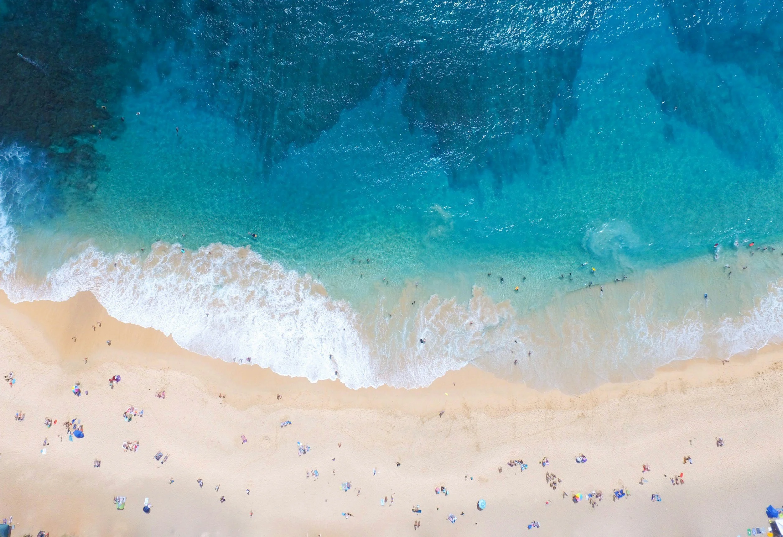 Aerial view of a beach with light-colored sand, colorful umbrellas, and people near the shoreline, with ocean waves crashing onto the beach.