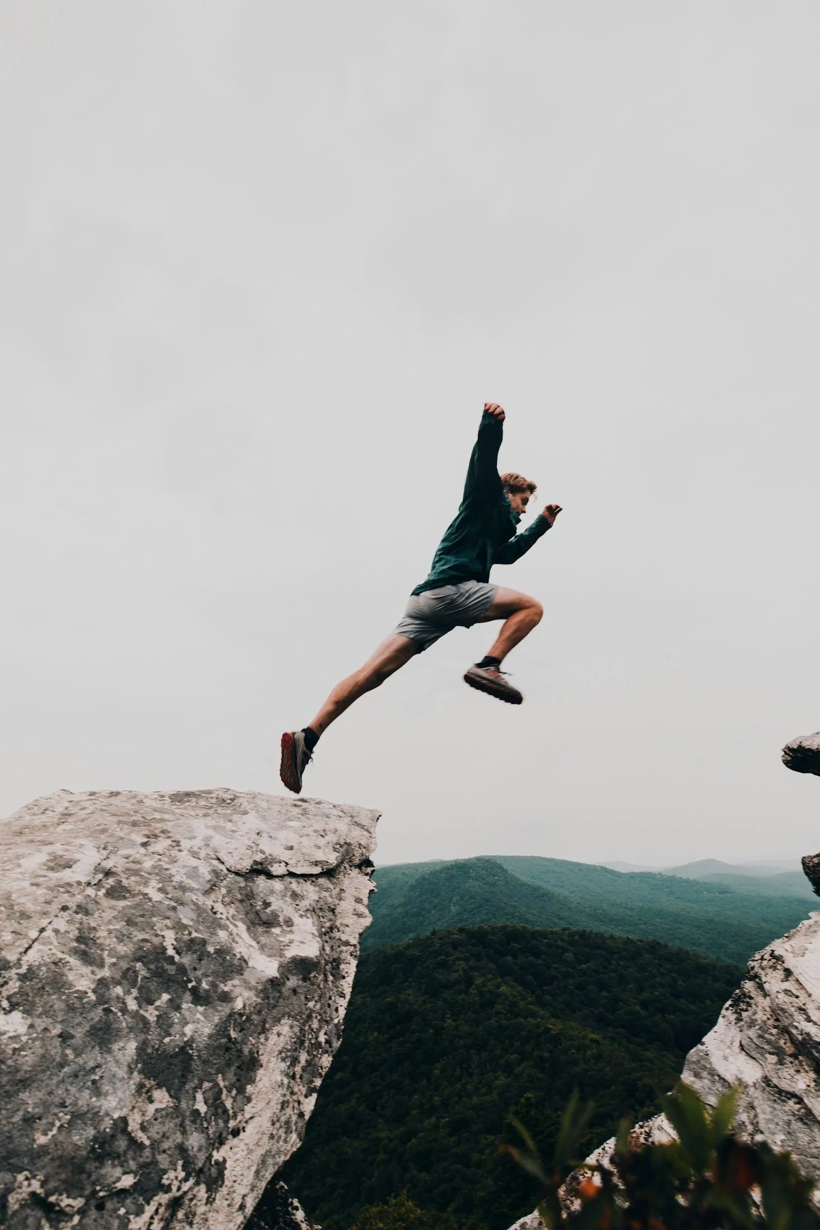 A man in sportswear leaps across a gap between rocky cliffs in a mountainous landscape under an overcast sky.