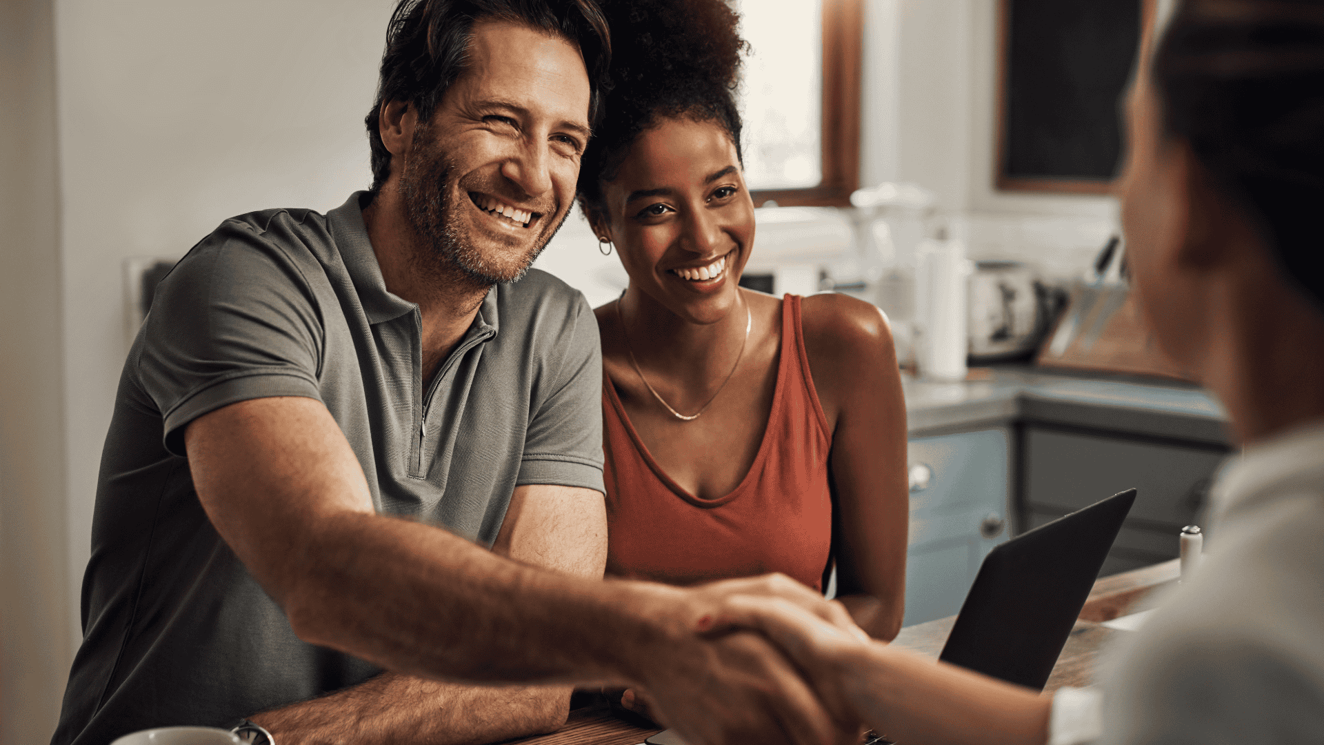 A man and woman smiling and shaking hands with a woman across a table in a kitchen.