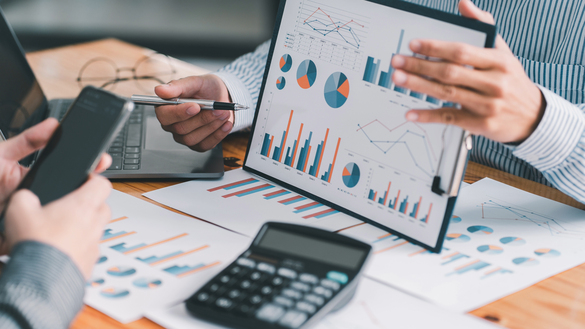 Business meeting analyzing graphs and charts on a tablet, with documents, calculator, laptop, and smartphones on a wooden table.