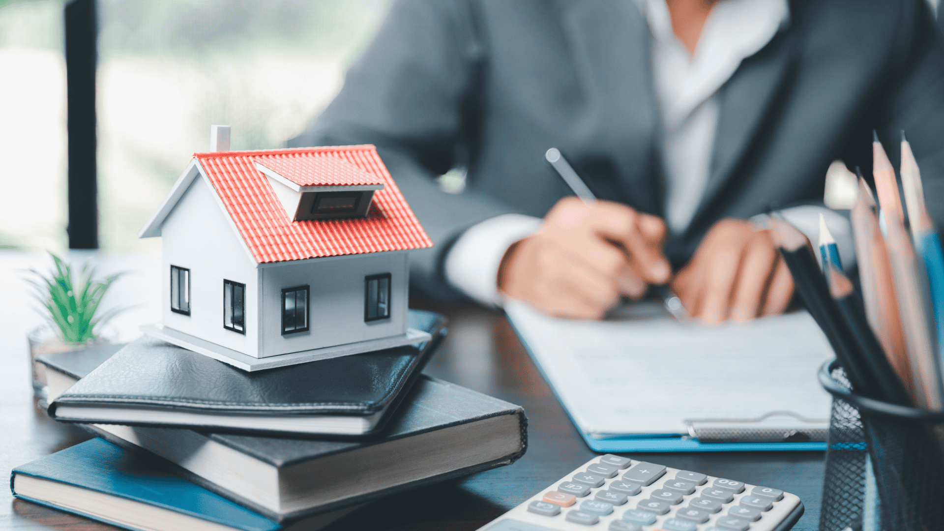A desk with a small model house on top of two closed books, a calculator, a potted plant, and a container with pens. A person in a suit is writing on a document in the background.