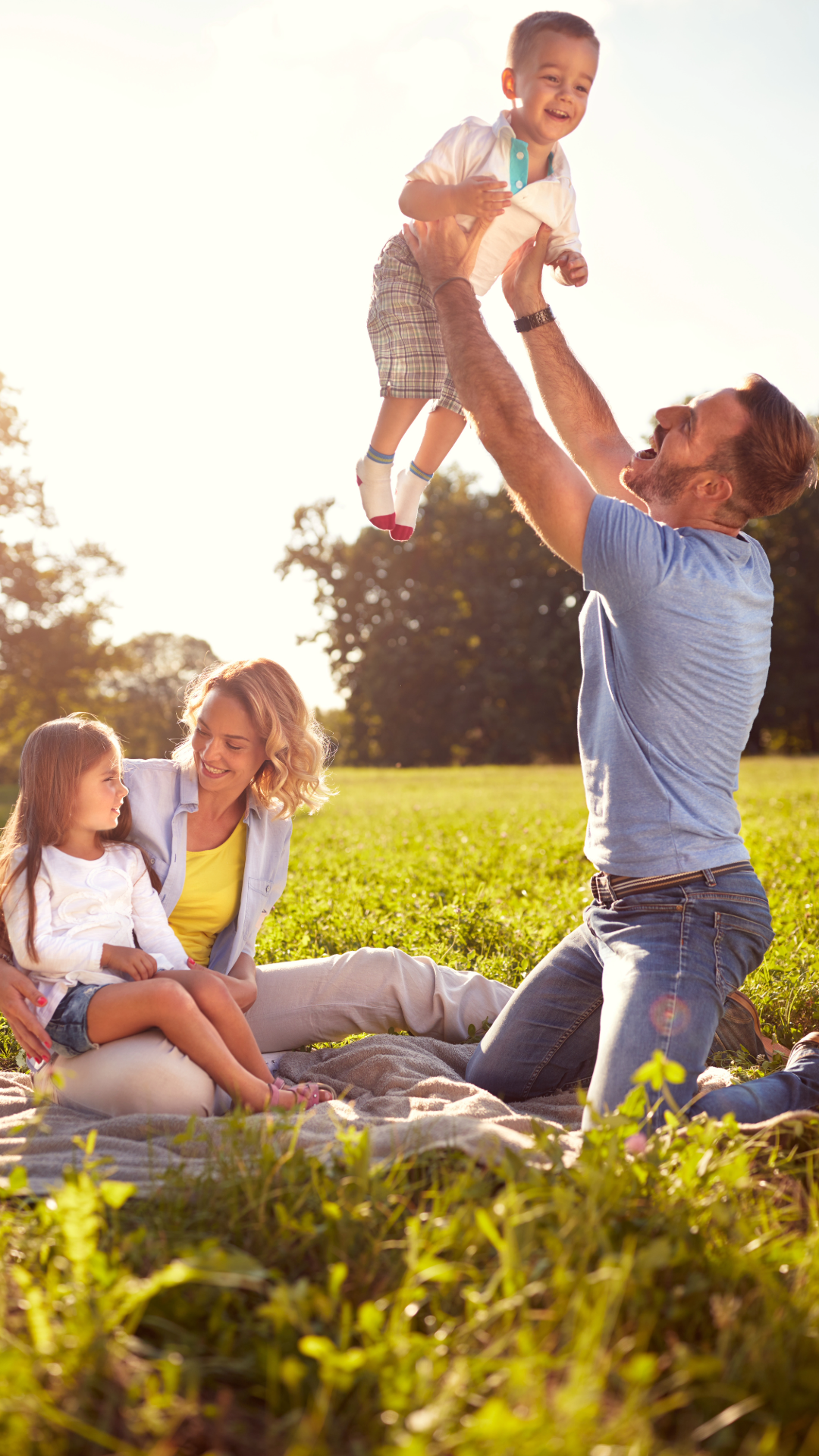 A family enjoying a sunny day outdoors. A man lifts a young boy into the air, while a woman and a girl sit on a blanket, smiling and watching.