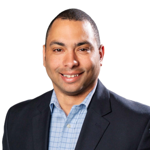 A smiling man in a dark suit and light blue shirt against a plain white background.