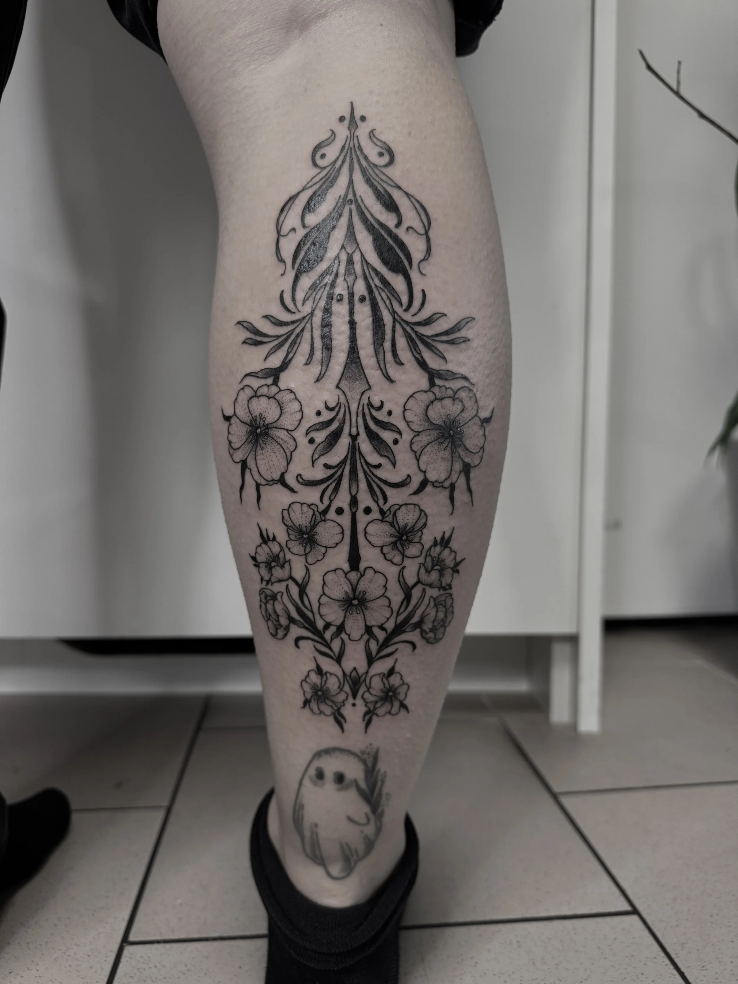 Schwarzes, detailliertes Tattoo eines Blumenmusters und eines kleinen symbolhaften Gesichts auf dem Bein einer Person.