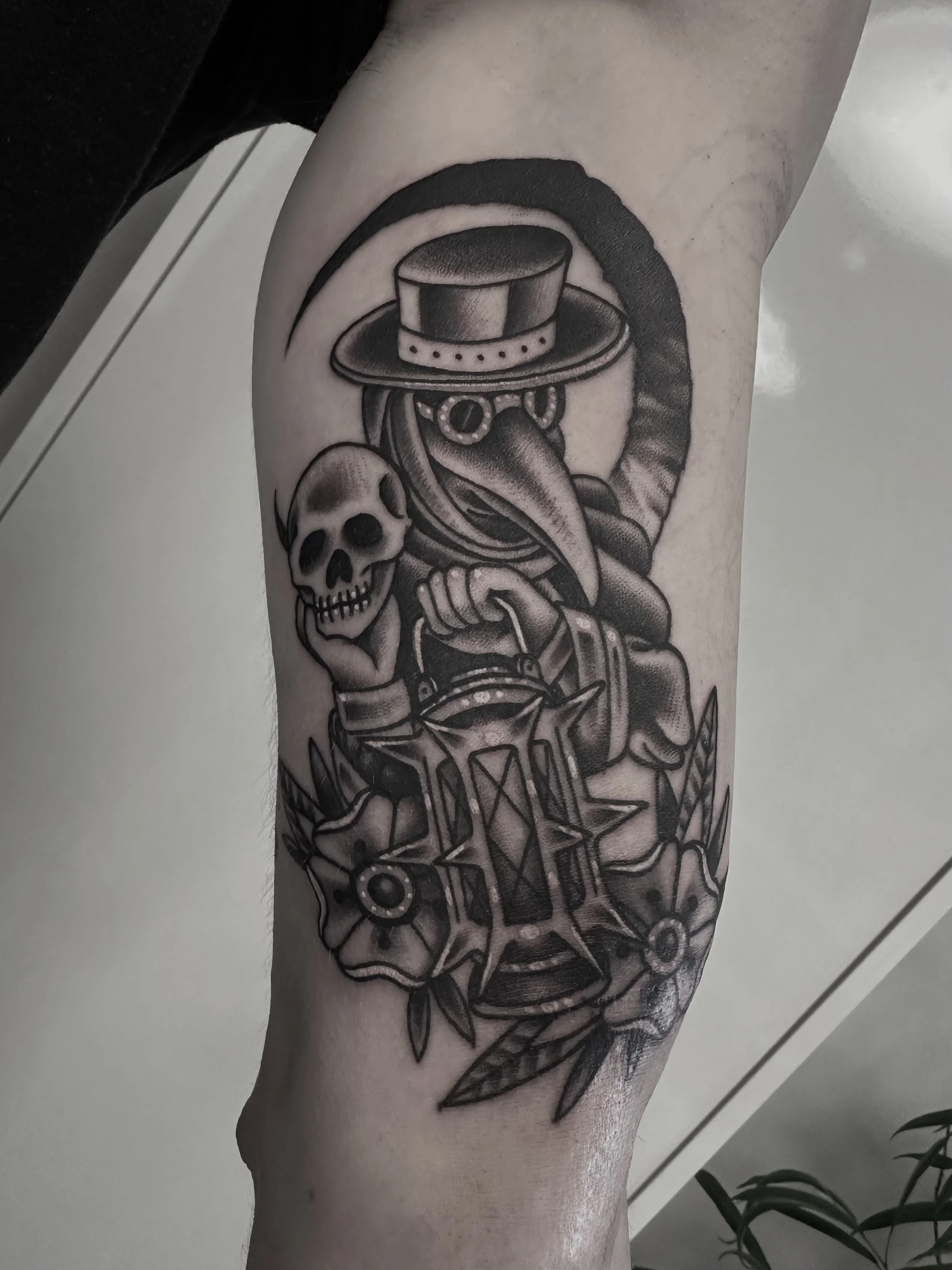 Schwarze Tätowierung eines Raben mit einem Zylinderhut, einer Brille, einer Lanzette und einer Hand, die eine Totenschädel hält, umgeben von Blumen auf dem Oberschenkel.